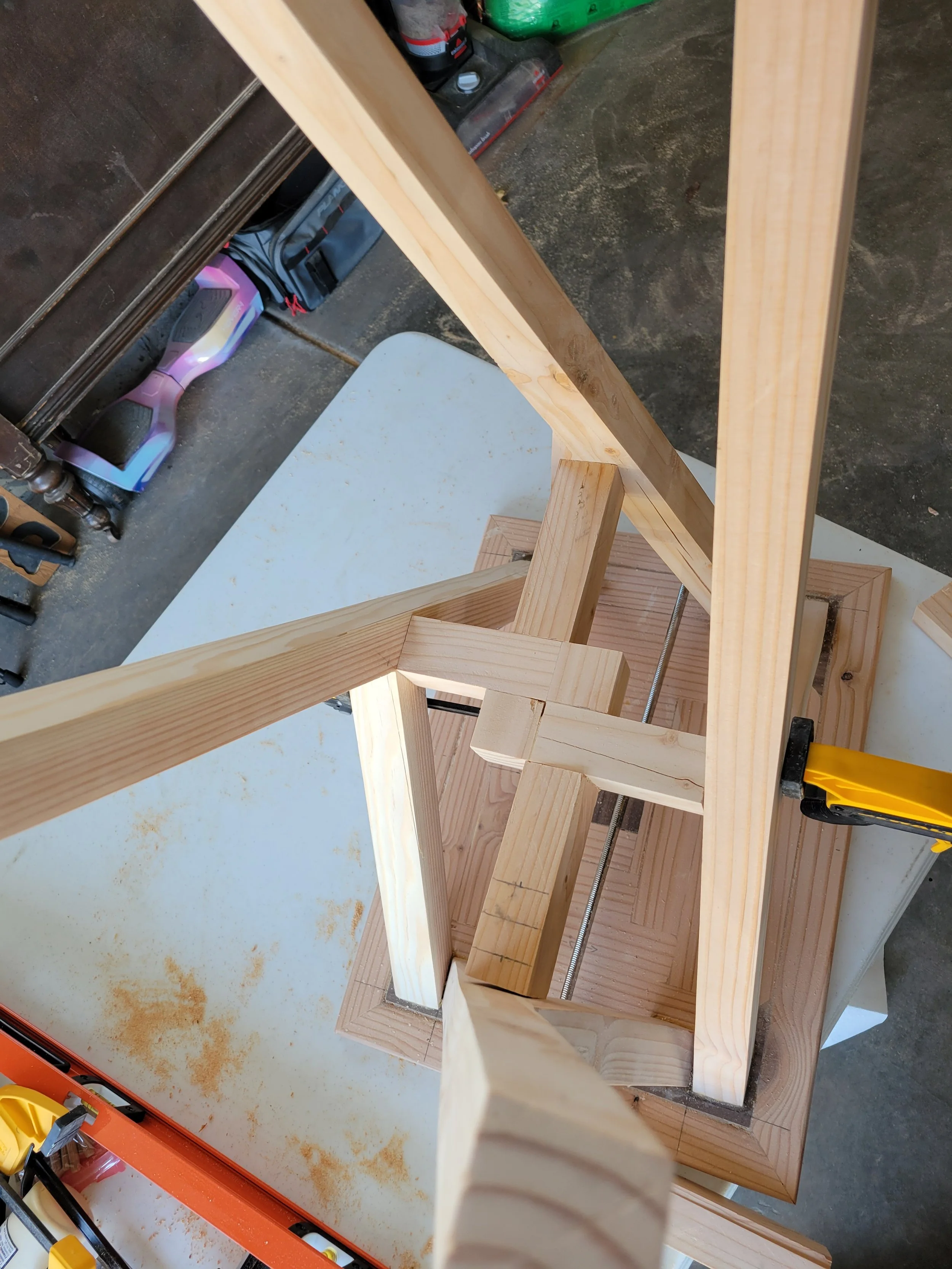 A woodworking project in progress showing wooden frame pieces clamped together on a workbench, surrounded by tools and equipment in a workshop.