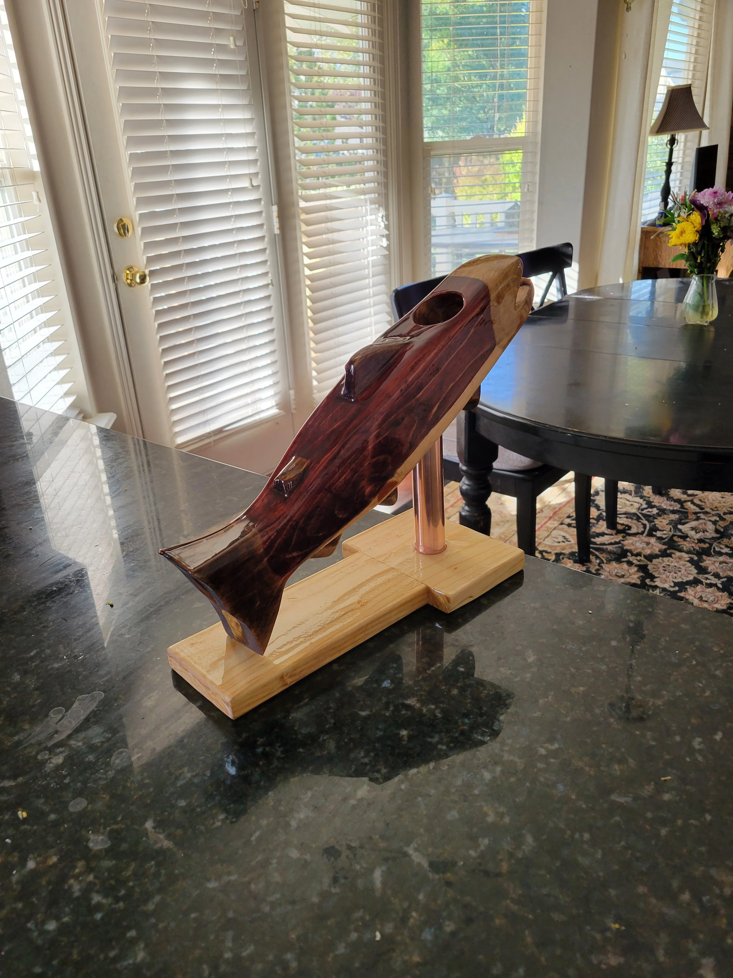 Wooden shark sculpture on display in a kitchen or dining area with large windows and blinds, a dark table with chairs, and a vase of colorful flowers.