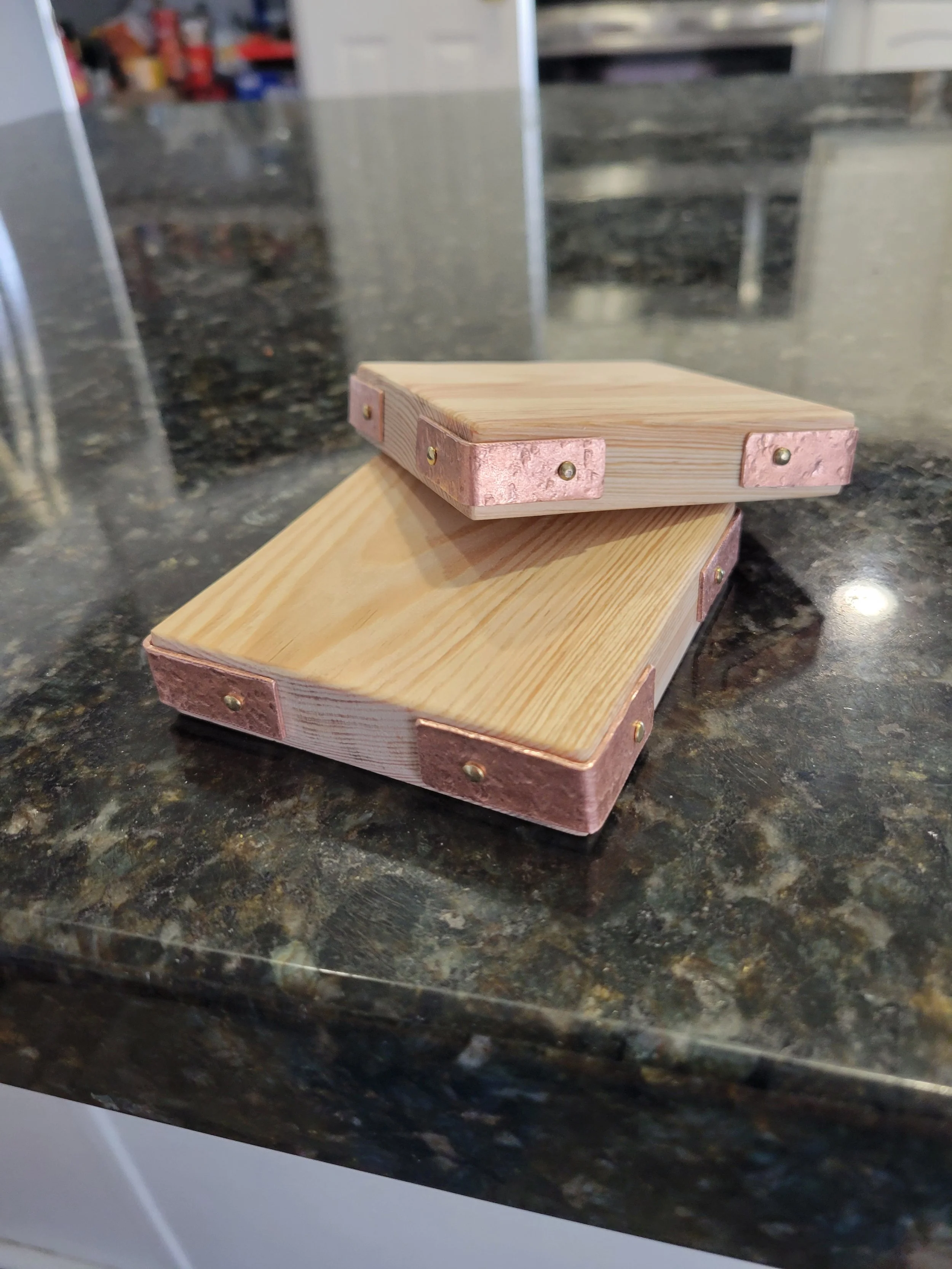 Two stacked wooden cutting boards with copper corner protectors and brass nails on a dark granite countertop.