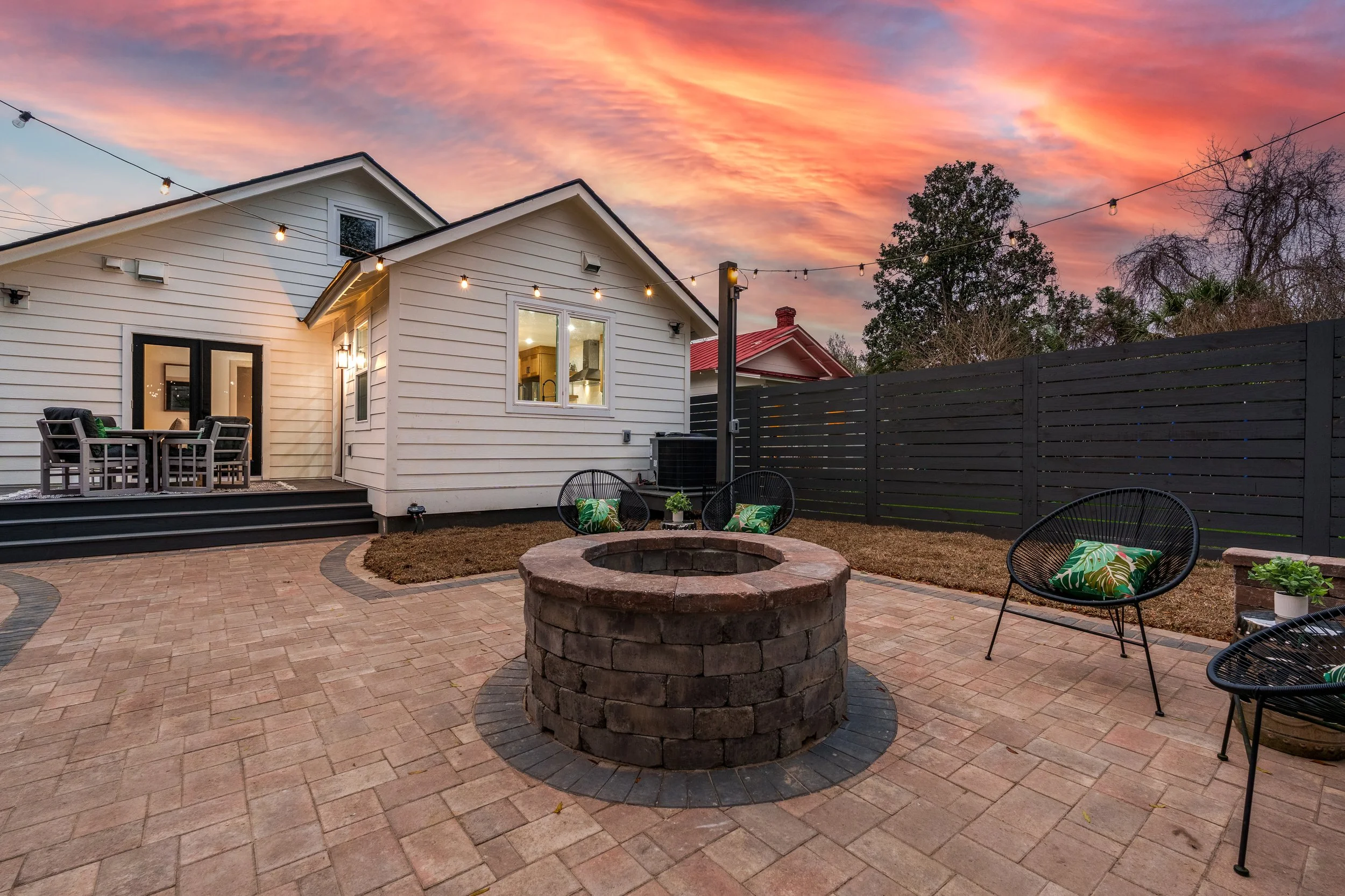 Backyard patio at sunset with brick fire pit, black outdoor chairs with green leaf pillows, wooden deck with dining table, string lights, white house with deck, and trees with colorful sky.
