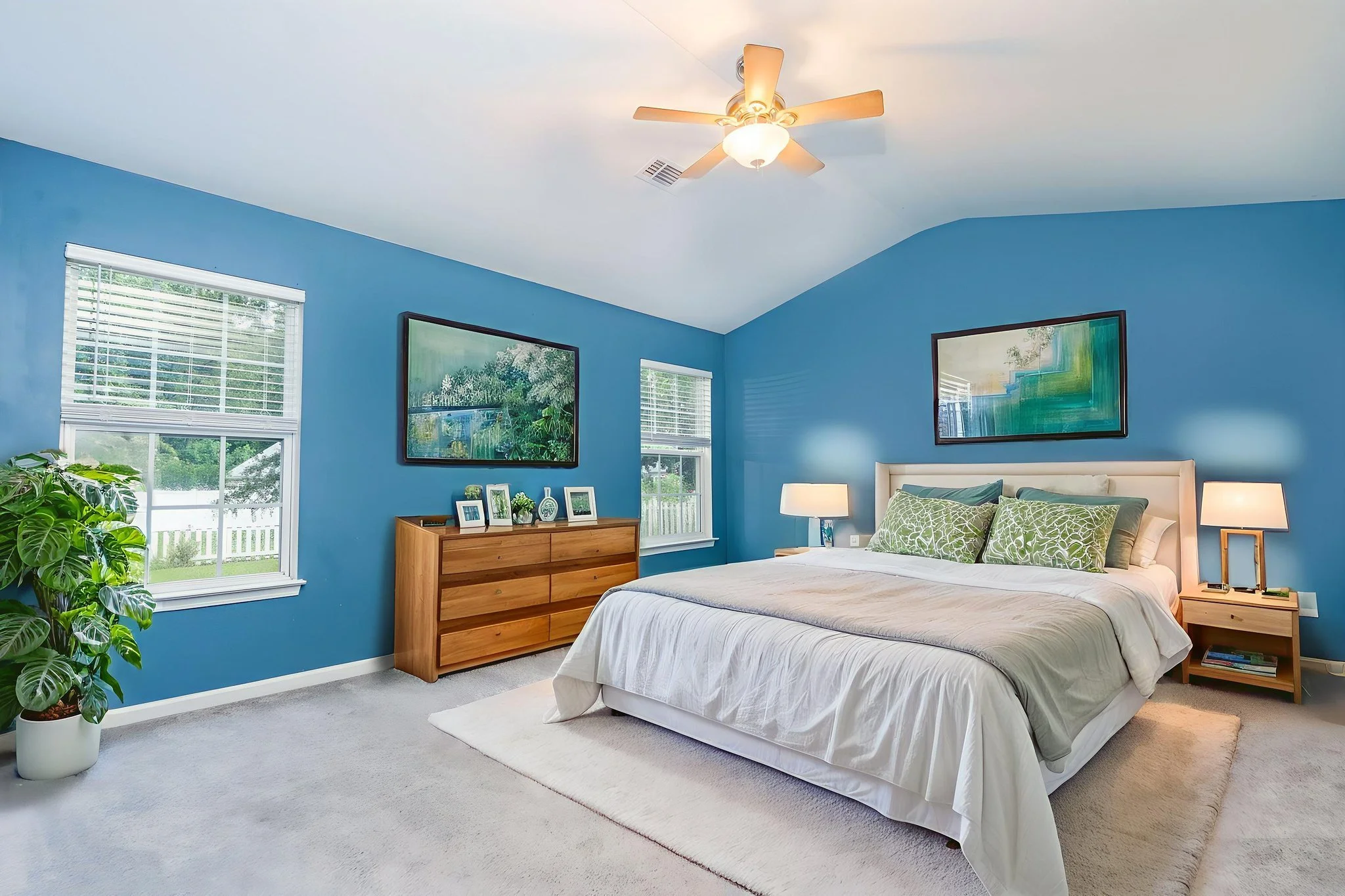 A bedroom with blue walls, a white bed with green pillows, wooden nightstands with lamps, a dresser with framed pictures, windows with blinds, a ceiling fan, and a potted plant.