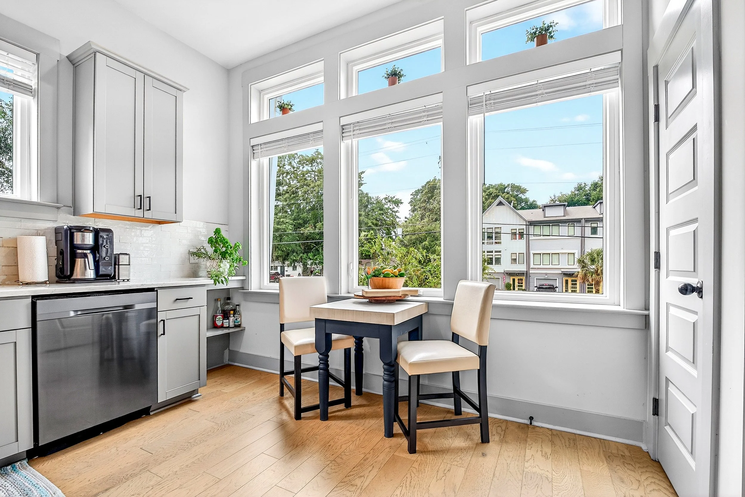 Bright kitchen with large windows, small table, two chairs, cabinets, countertop, and a view of neighboring houses and trees outside.