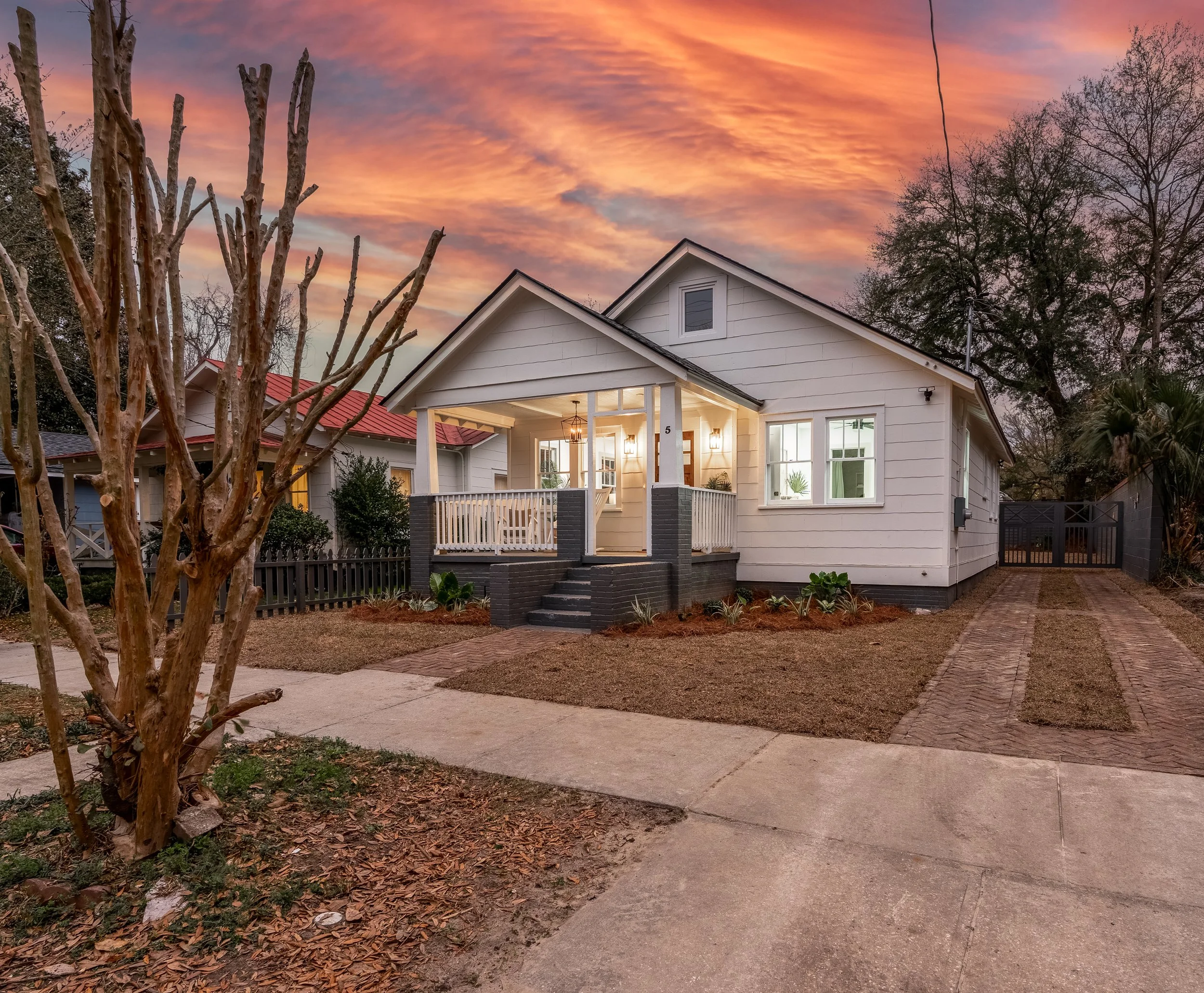 A cozy white house with a front porch illuminated at sunset, with a brick and concrete driveway to the right and leafless tree in the front yard.