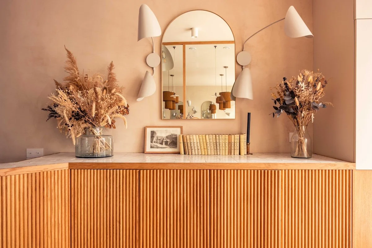 A modern wooden sideboard with a marble top, decorated with two vases of dried flowers, a framed photo, a row of vintage books, and a candle holder. A large arched mirror and two wall-mounted lamps are above the sideboard, with warm lighting and neutral-colored walls creating a cozy atmosphere.