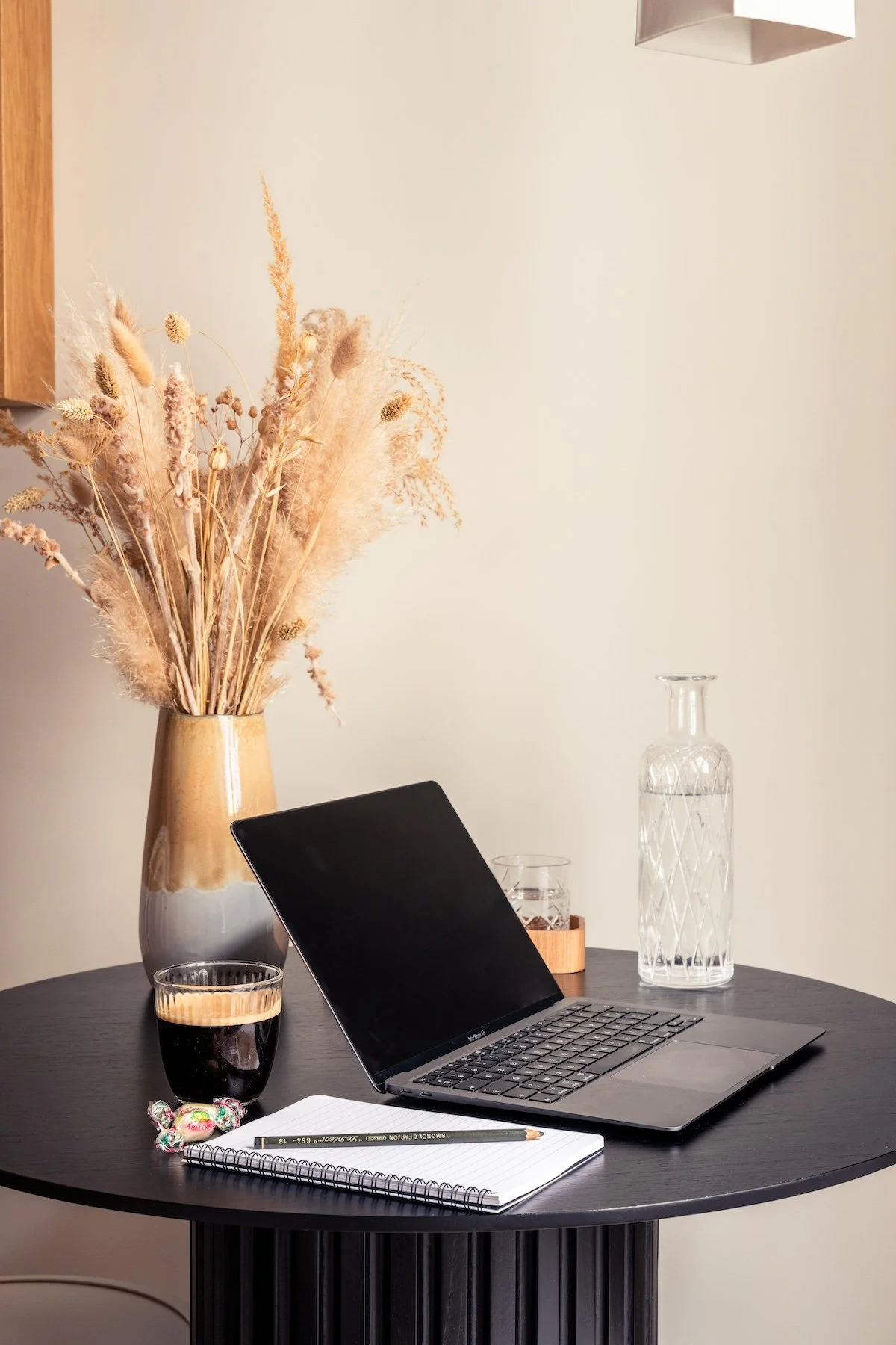 A round black table with a laptop, a glass of dark beverage, a notepad with a pencil, a large clear glass water bottle, a small wooden tray with a glass, a vase of dried flowers, and wrapped candies.