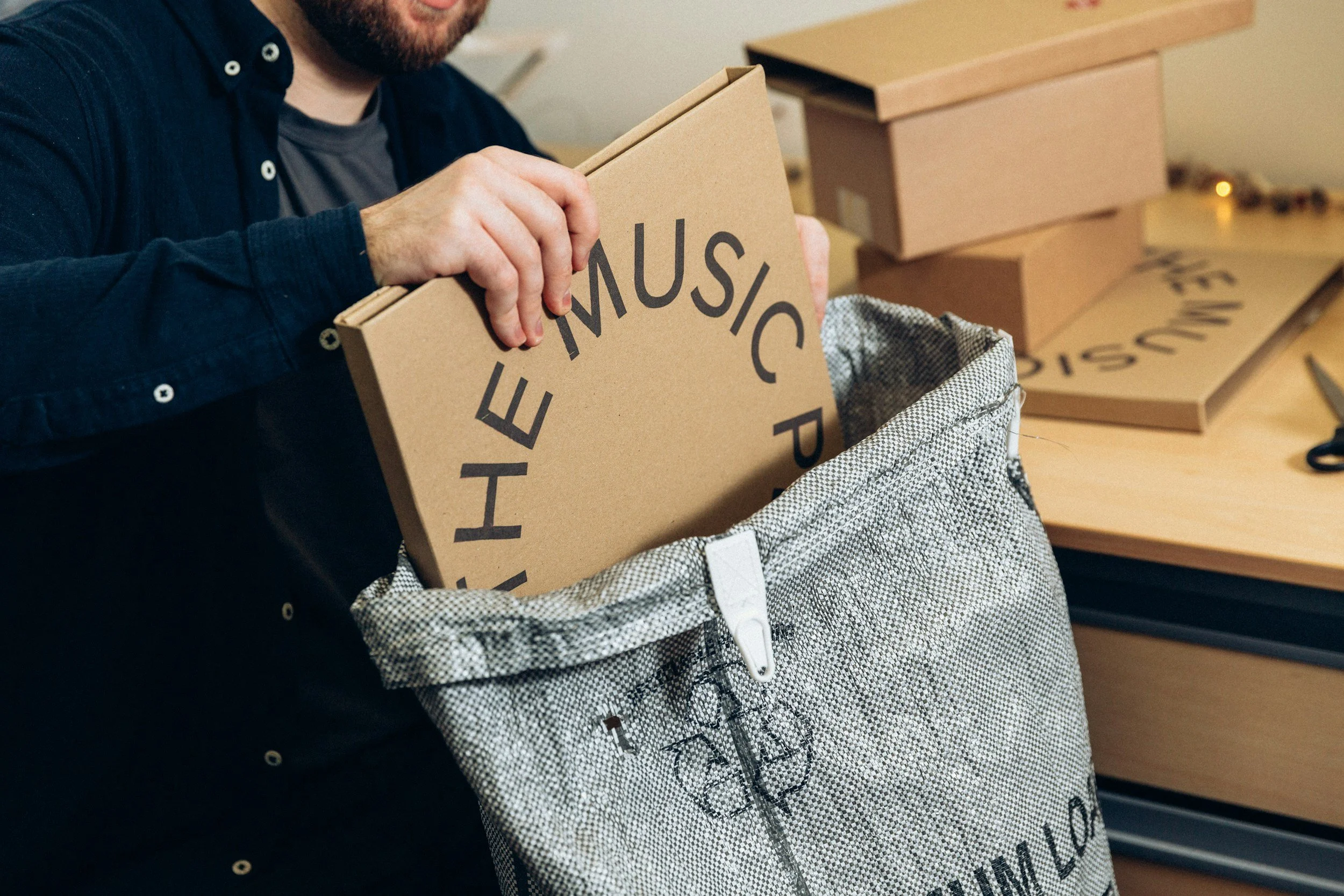 Person packing a cardboard sign that says "THE MUSIC" into a gray fabric bag on a wooden table.