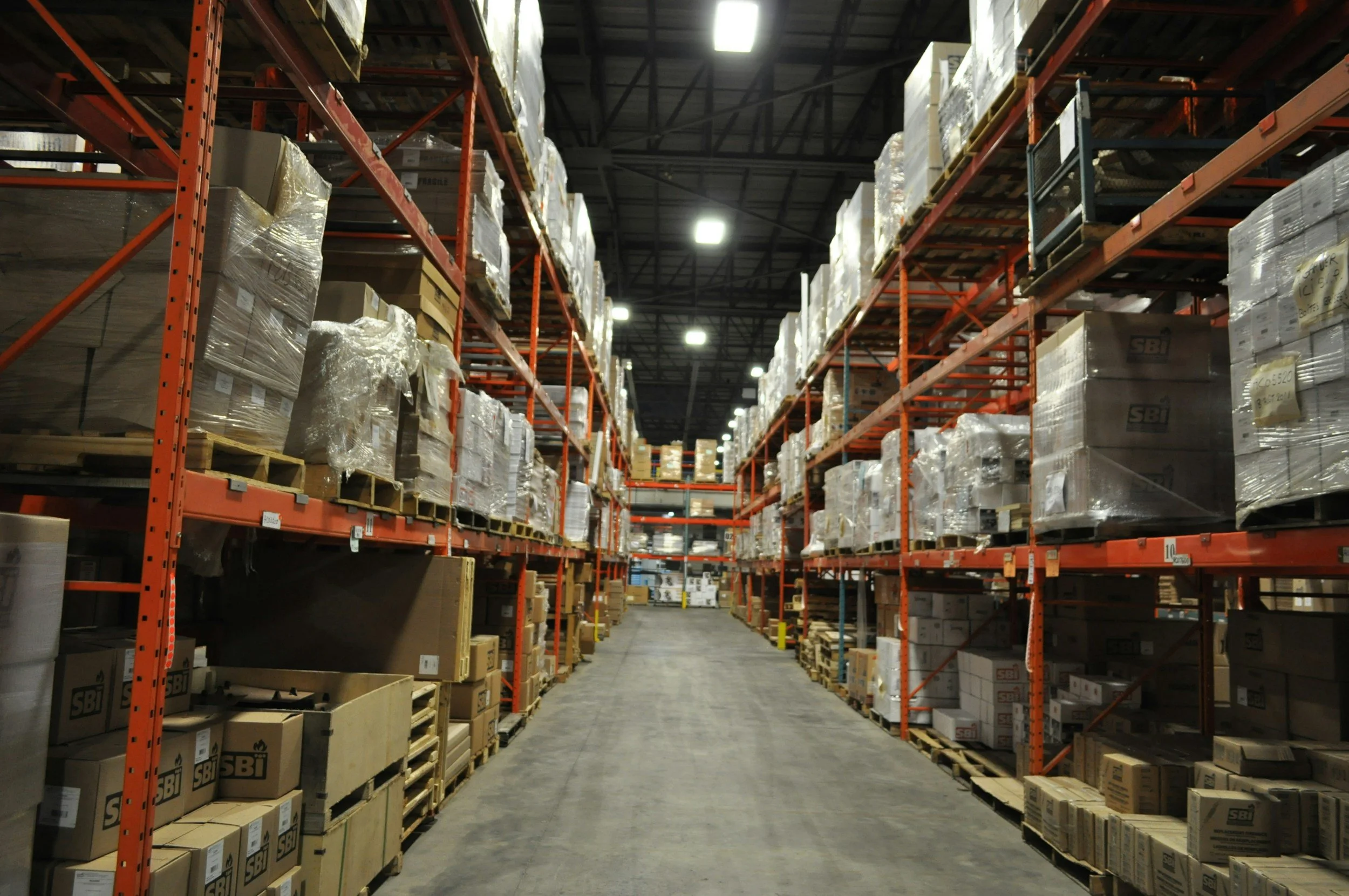 A wide aisle in a warehouse with tall orange metal shelving units on both sides, filled with stacked cardboard boxes wrapped in plastic, some on wooden pallets, and fluorescent ceiling lights overhead.