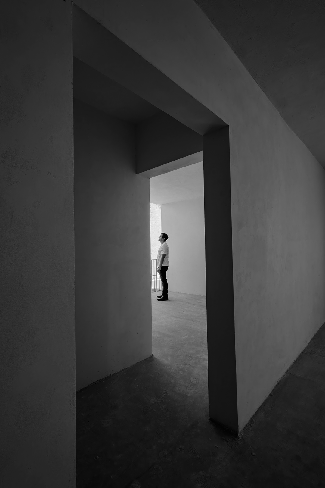 Un hombre de pie en un espacio arquitectónico con paredes de hormigón en blanco y negro, observando hacia adelante desde una área abierta con barandilla.