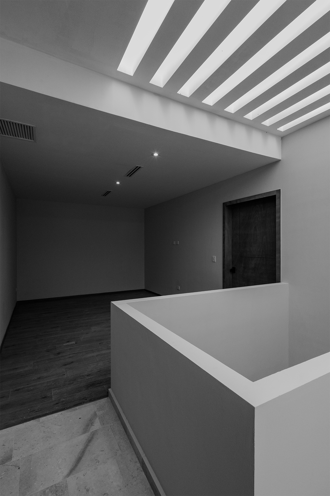 Interior de una casa moderna con techos altos y luz natural, paredes blancas, piso de madera y puerta de madera oscura.