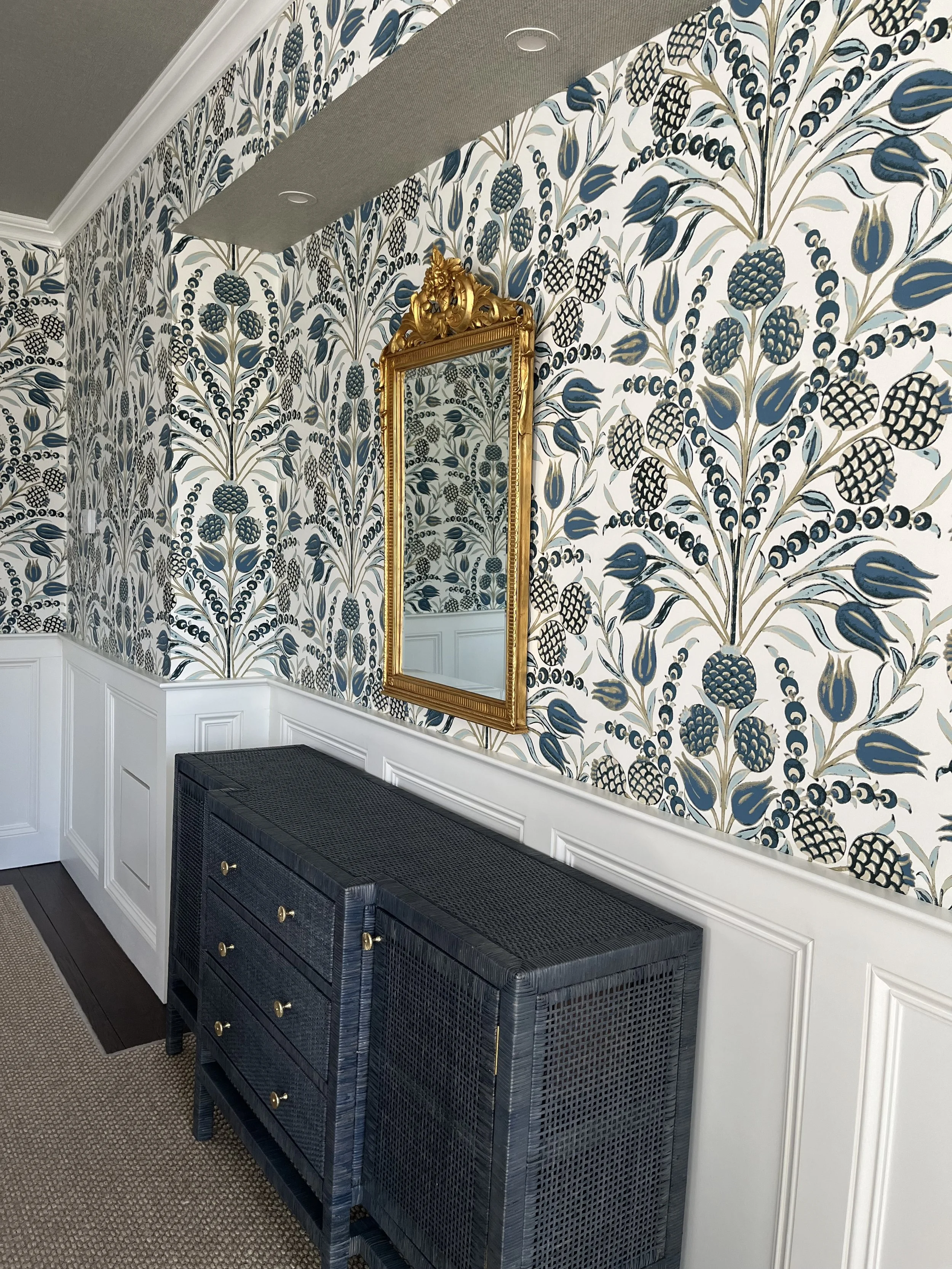 A living room wall with blue, beige, and white patterned wallpaper, a gold-framed mirror, a white wainscot, a woven black storage bench, and a carpeted floor.
