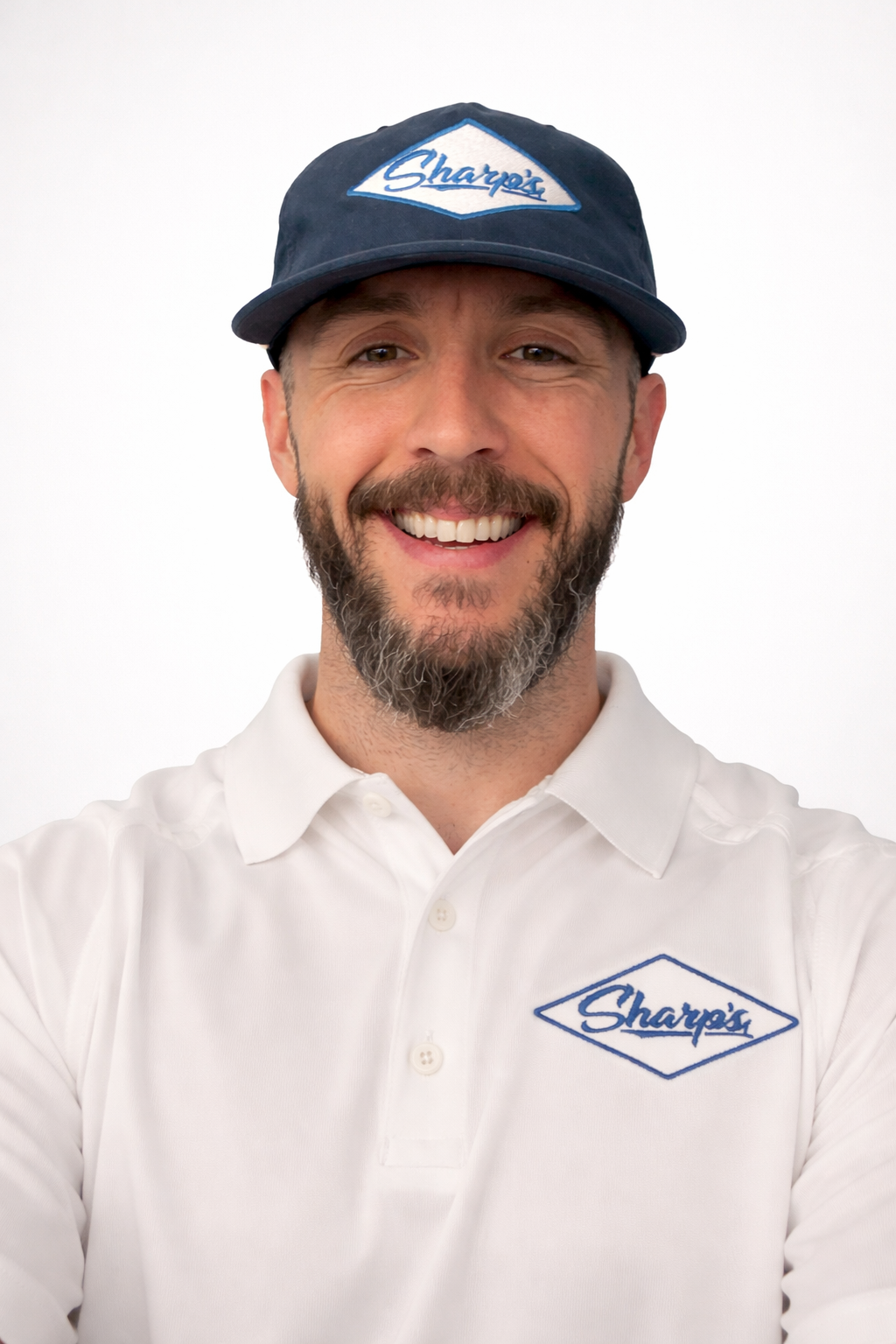 A man with a beard wearing a white polo shirt and a blue cap with the Sharpi's logo, smiling against a plain white background.