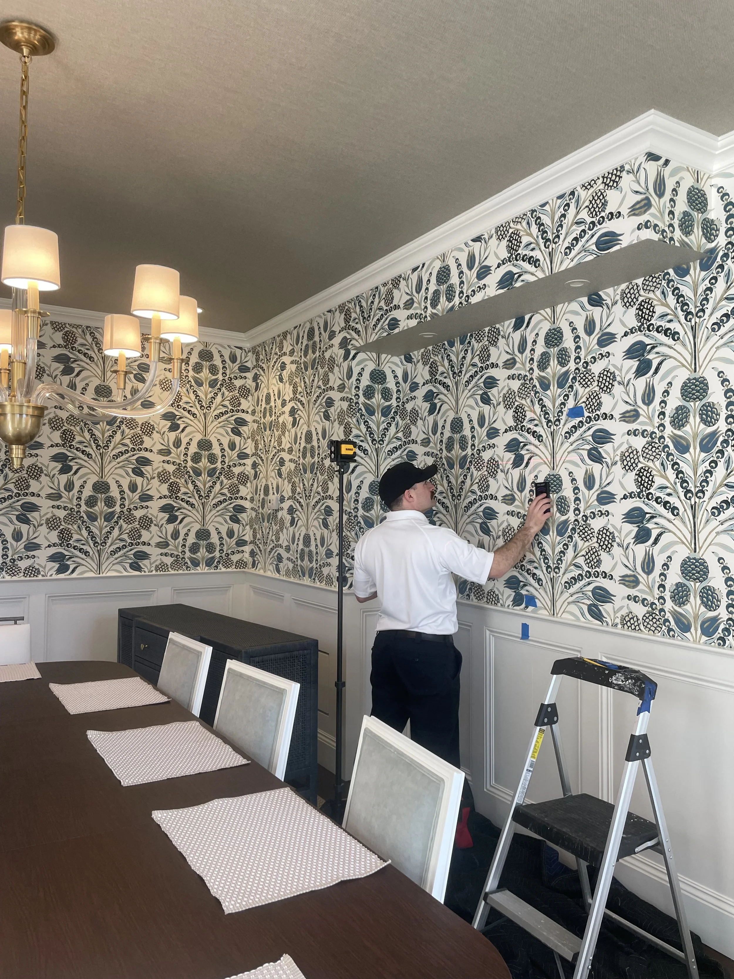 A person standing on a ladder, measuring or fixing wallpaper in a dining room with a large dining table and chairs, chandelier, and decorative wall trim.