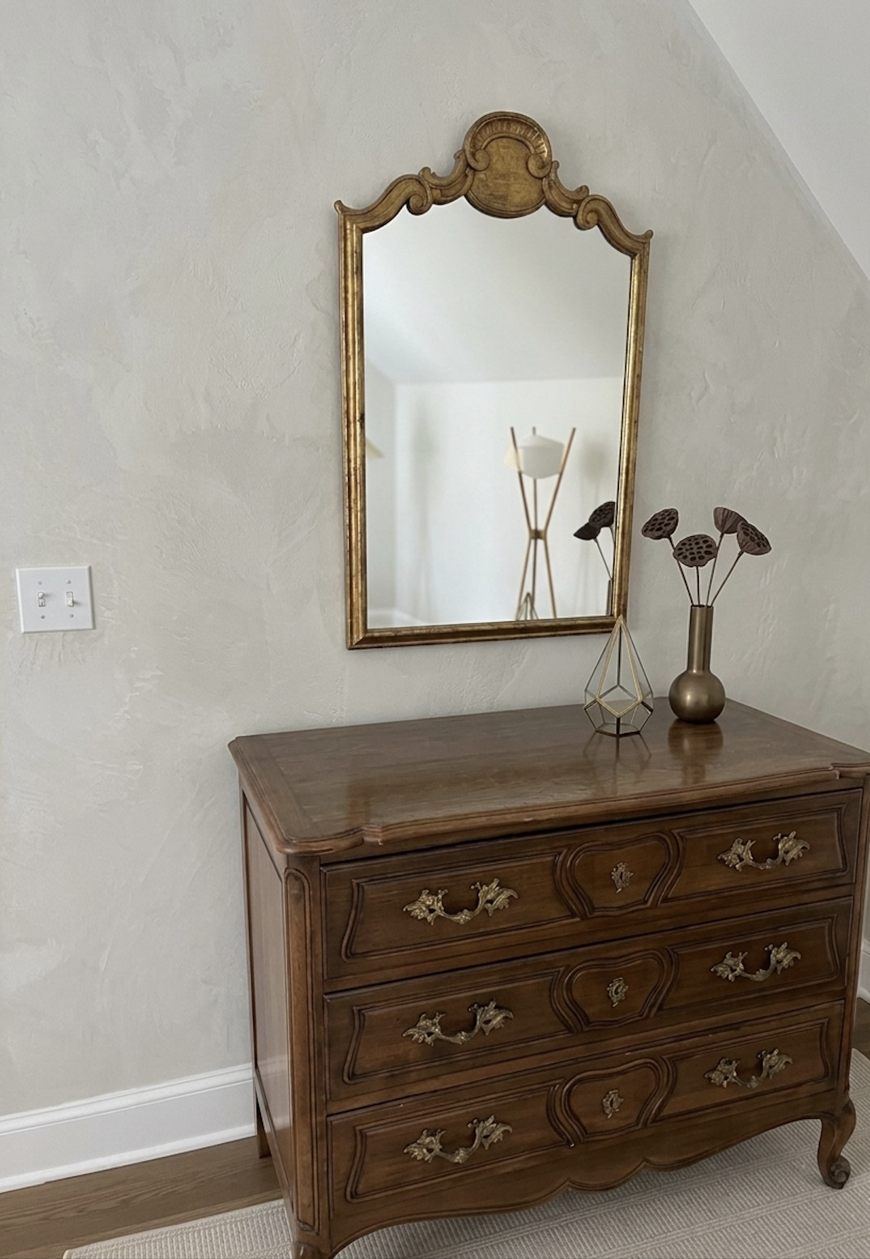 A vintage wooden dresser with ornate gold handles, topped with decorative vases and a geometric sculpture, reflected in a gold-framed mirror on a beige textured wall.