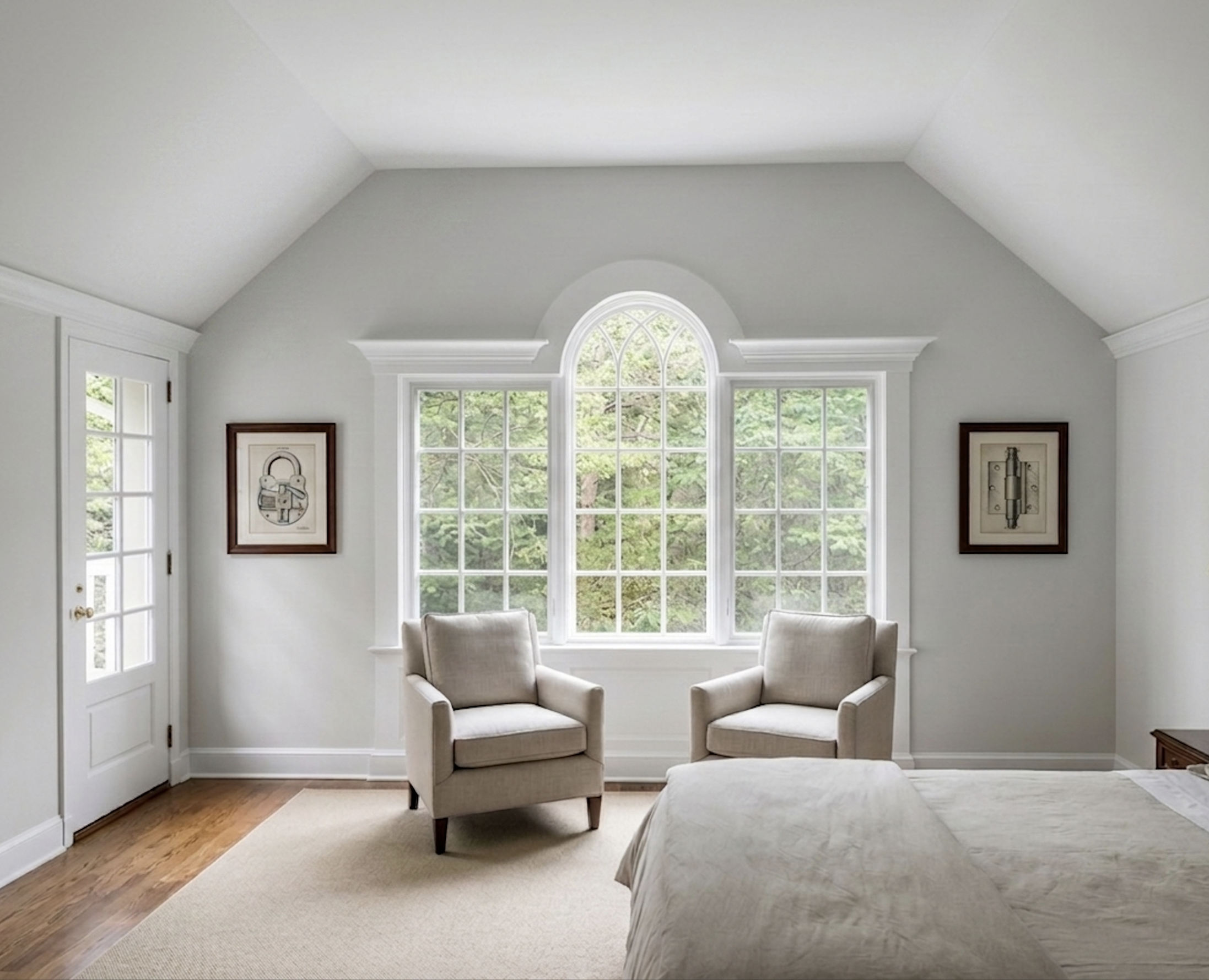 A well-lit living room with large, arched windows showing green trees outside. There are two beige upholstered armchairs, a bed, framed artwork on the walls, and a wooden side table.