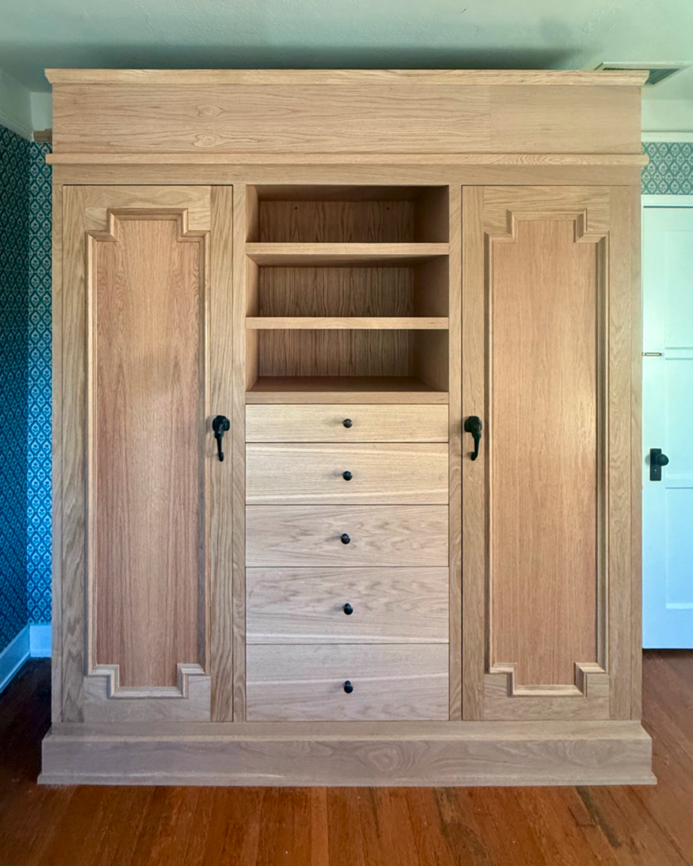 Natural wood wardrobe with two paneled doors, open shelves in the middle, and five drawers below, located on a wooden floor.