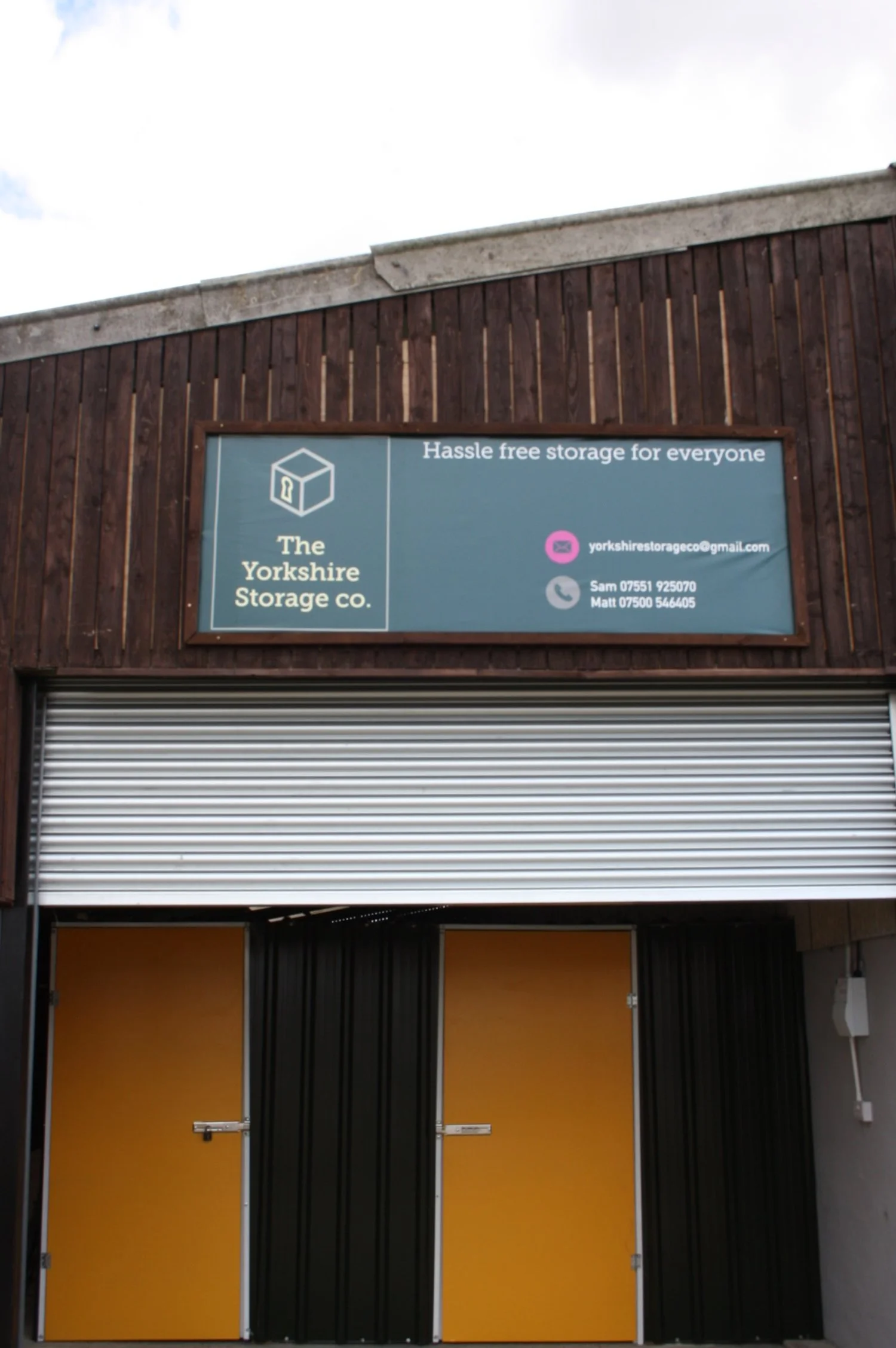 Sign for Yorkshire Storage Co. offering hassle-free storage, with contact details including email 'yorksharestorageco@gmail.com' and phone numbers for Sam and Matt, installed on a wooden building with a partially open metallic yellow roll-up door.