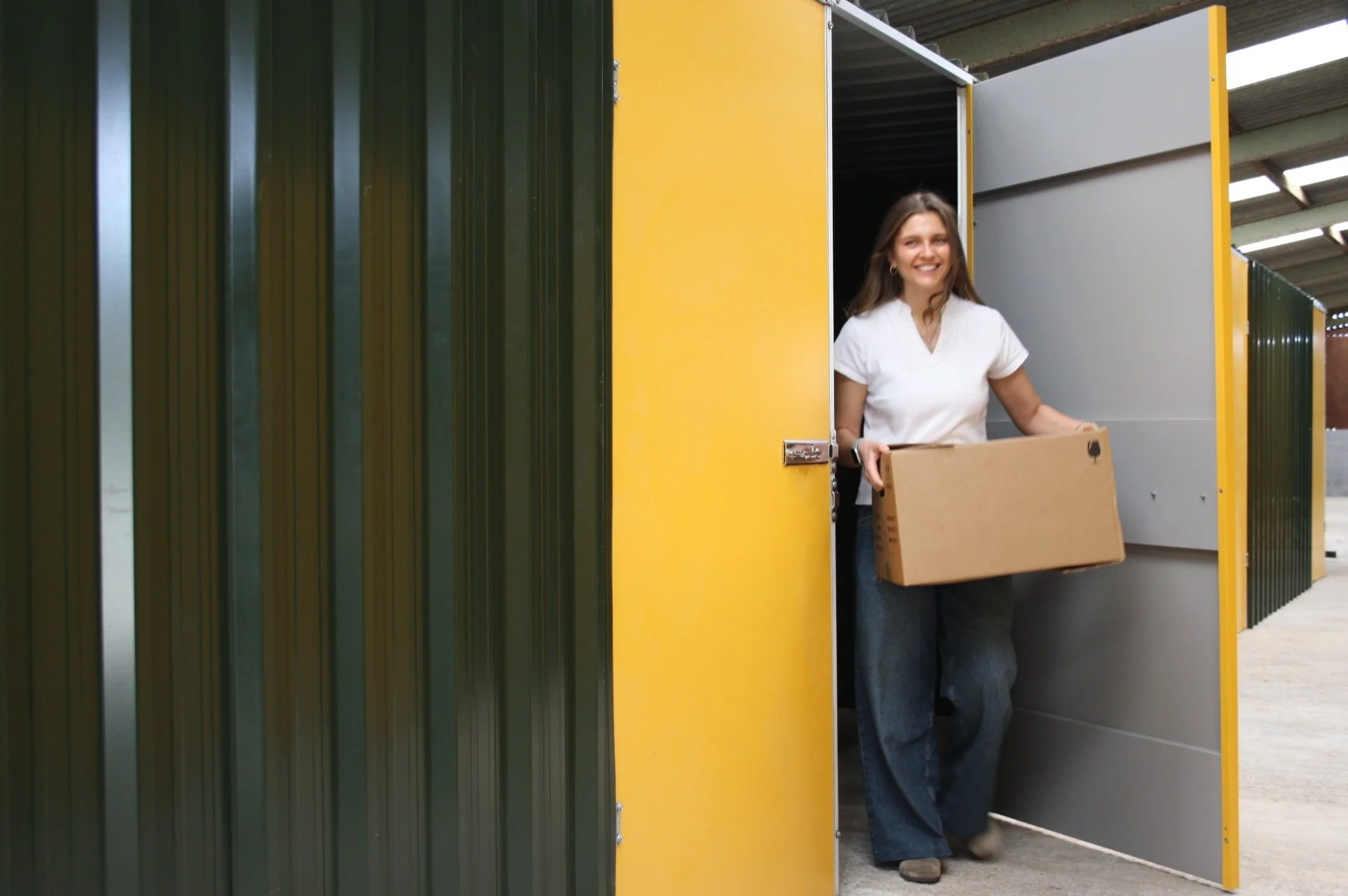 A woman walking out of a storage unit holding a cardboard box, smiling.