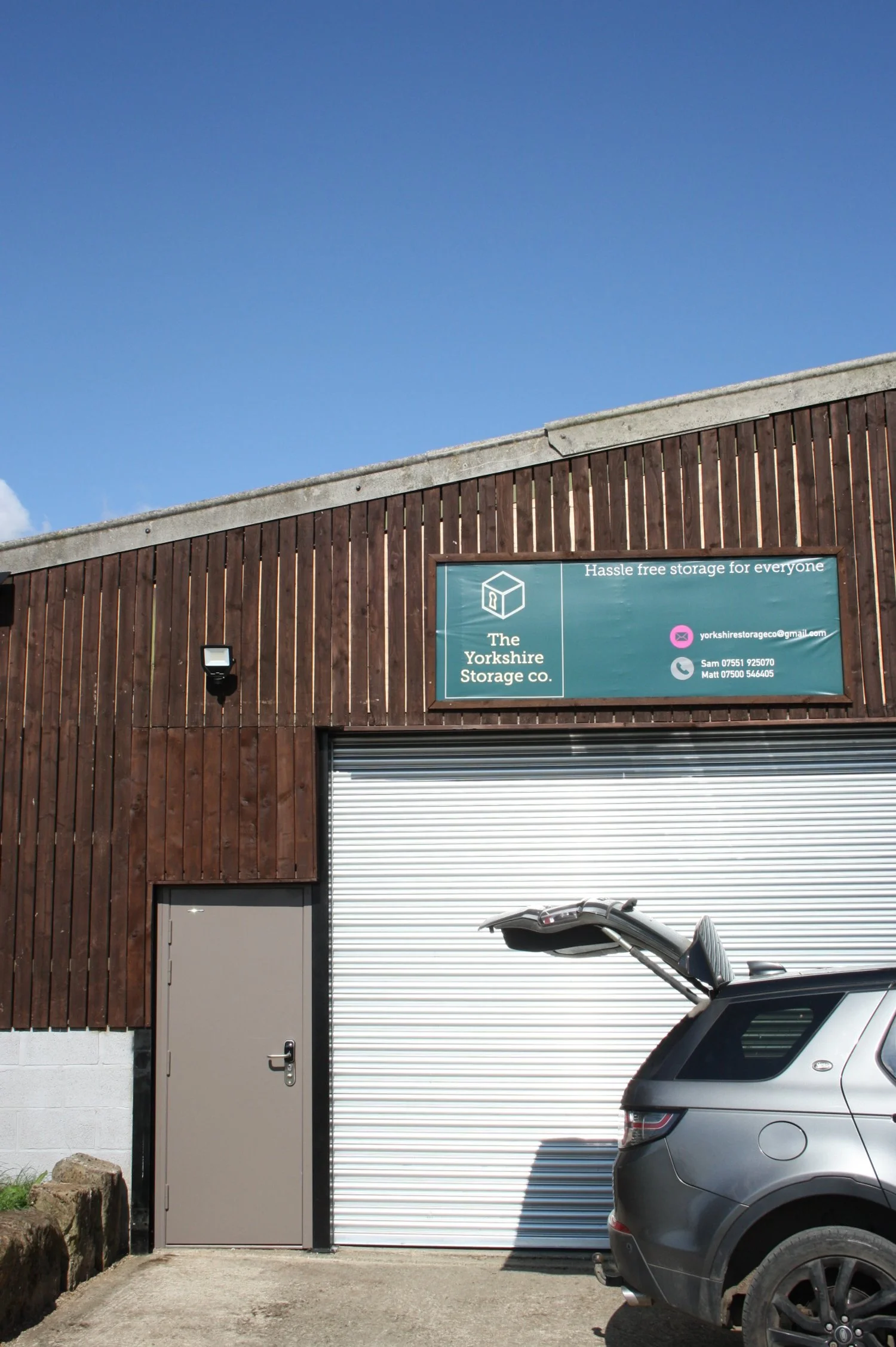 Exterior view of a storage facility building with a wooden facade, a grey door, a metal roller shutter, and a silver SUV with an open trunk. There is a sign on the building that reads 'The Yorkshire Storage co.' and offers hassle-free storage for everyone. The sky is clear and blue.