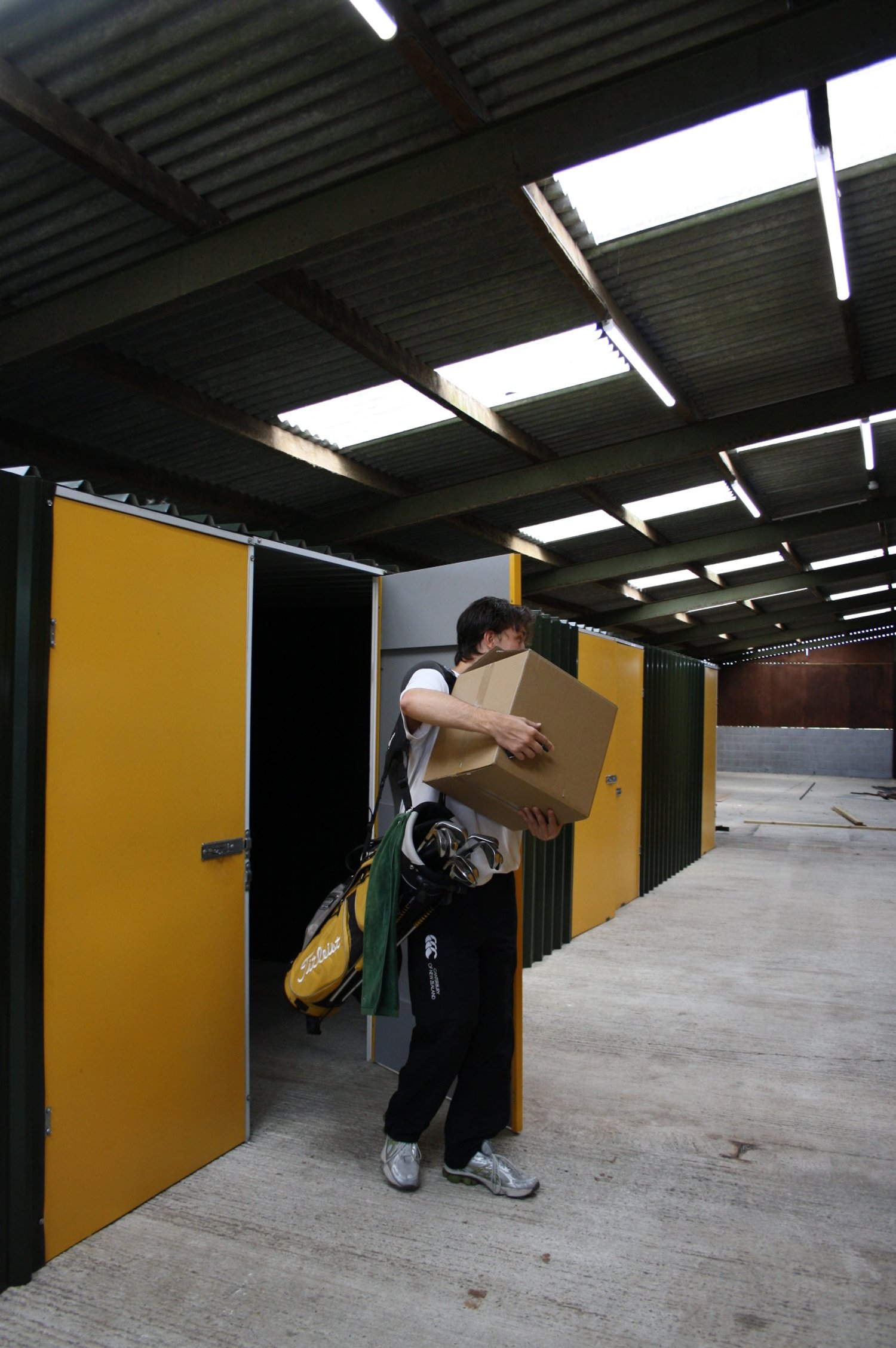 A person walking out of a locker room carrying a cardboard box, wearing athletic clothing and a golf bag with clubs over their shoulder, inside a large industrial or storage facility with a metal roof.