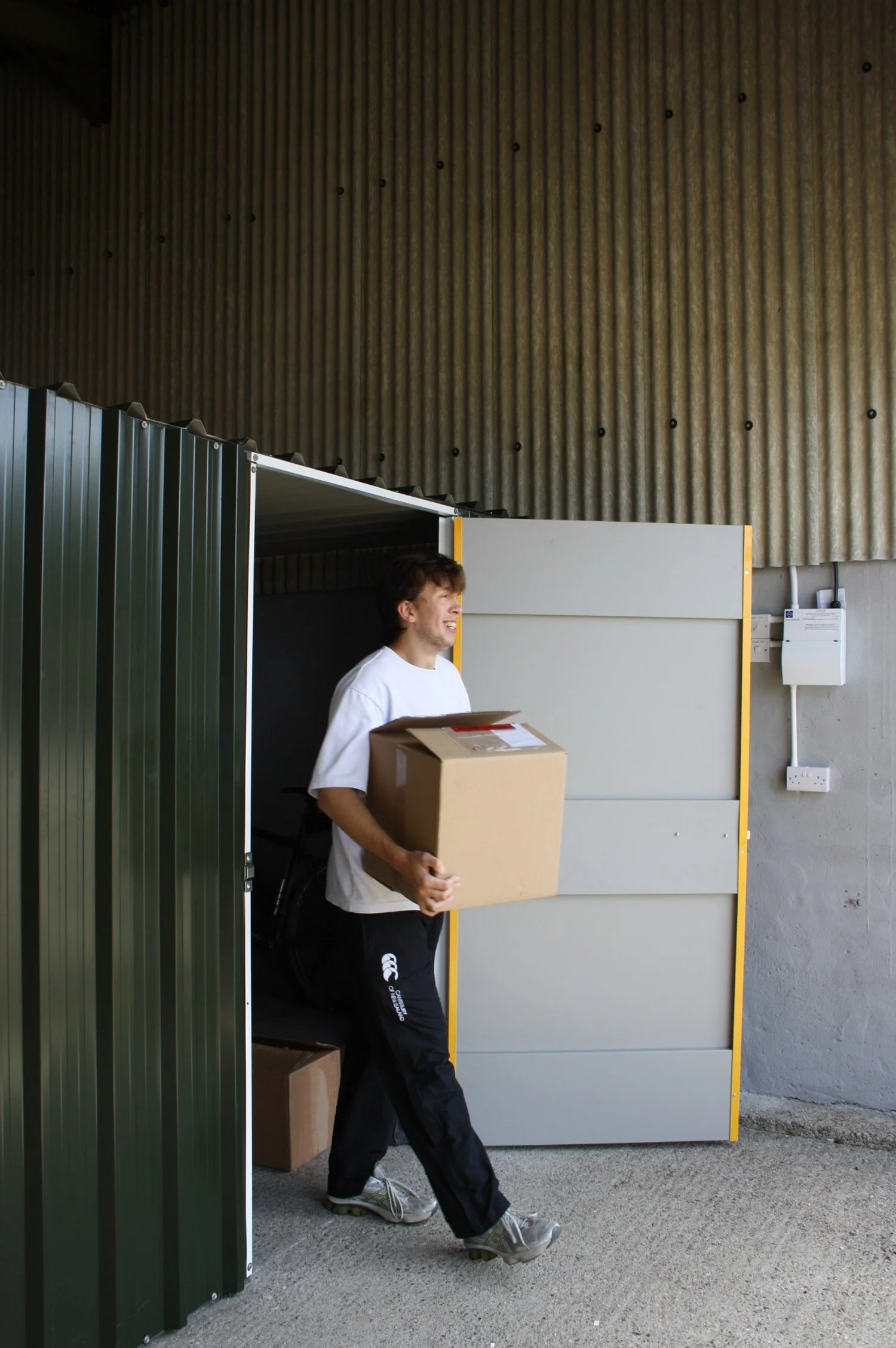 Young man smiling while carrying a cardboard box outside a storage unit or garage.