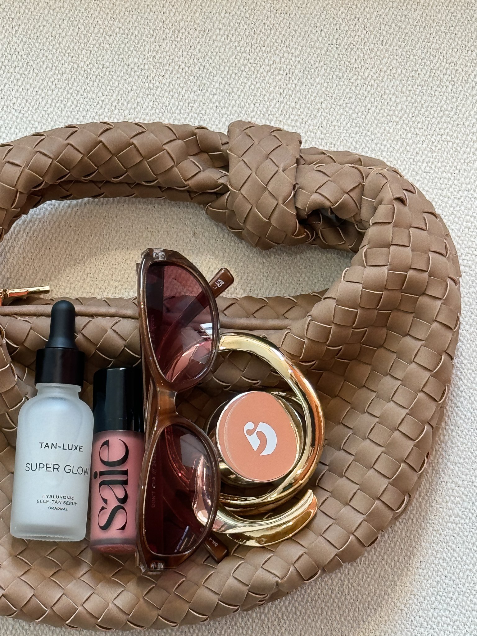 A woven tan handbag holding sunglasses, skincare serum, cheek blush, and makeup compact.