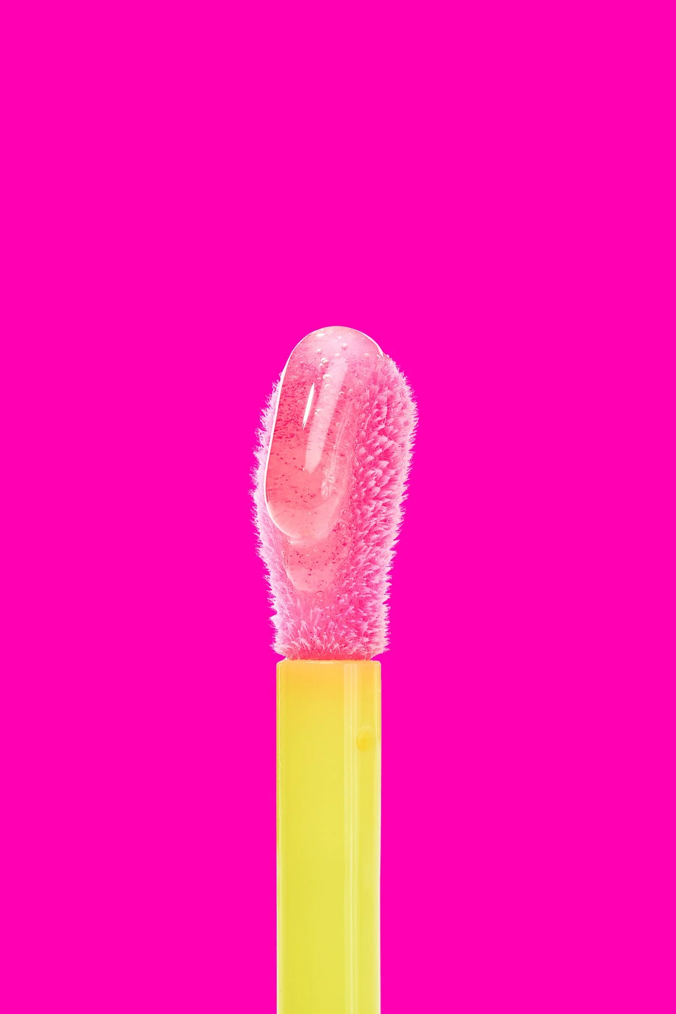 A close-up of a silicone makeup applicator with pink bristles and a clear gel-like substance on top, against a bright pink background.