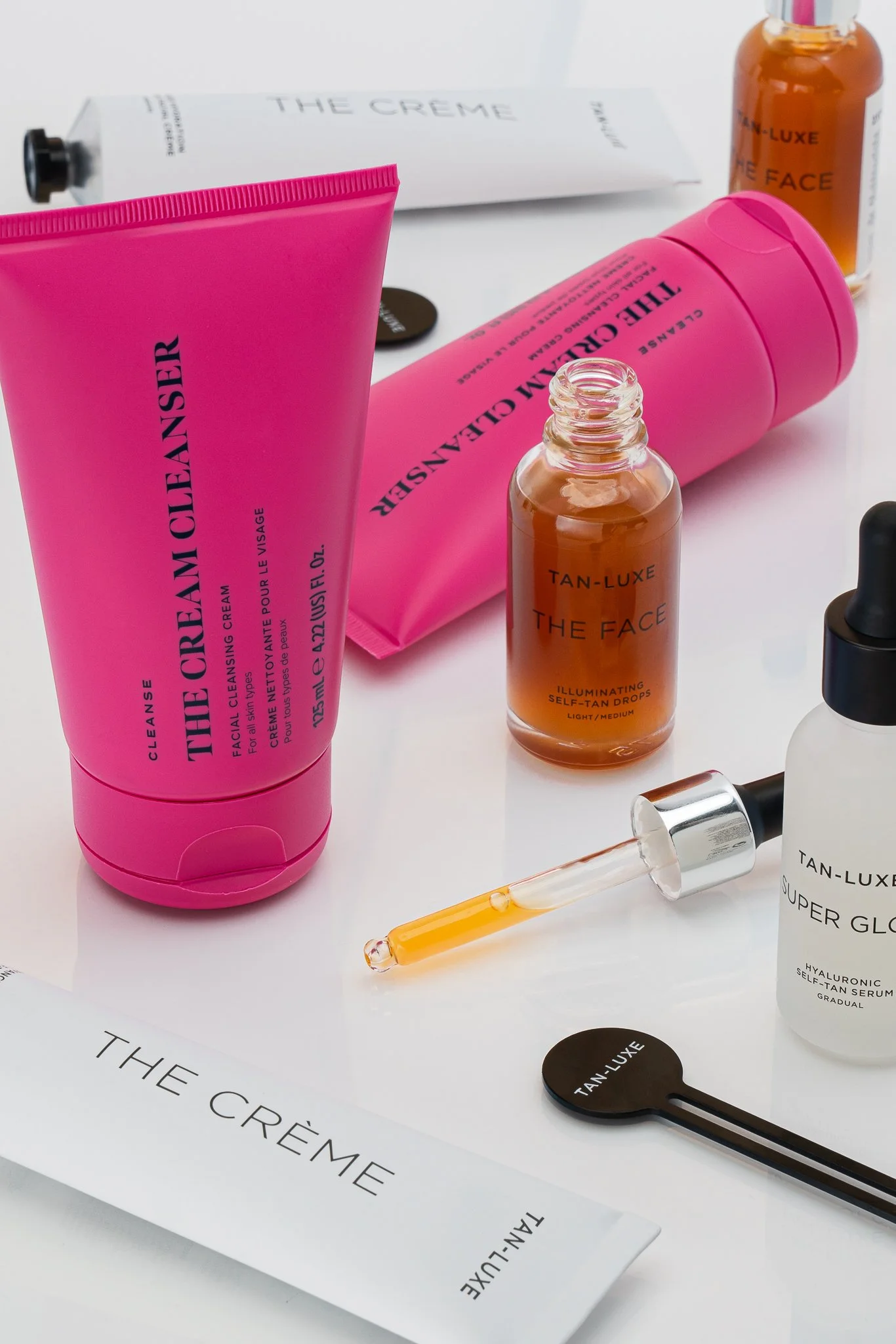 Skincare products including a pink tube labeled 'The Cream Cleanser,' a small amber bottle labeled 'Tan-Luxe The Face,' a white bottle labeled 'Super Glow,' a pink tube labeled 'The Cream,' a black applicator with 'Tan-Luxe' on it, and a glass droppe