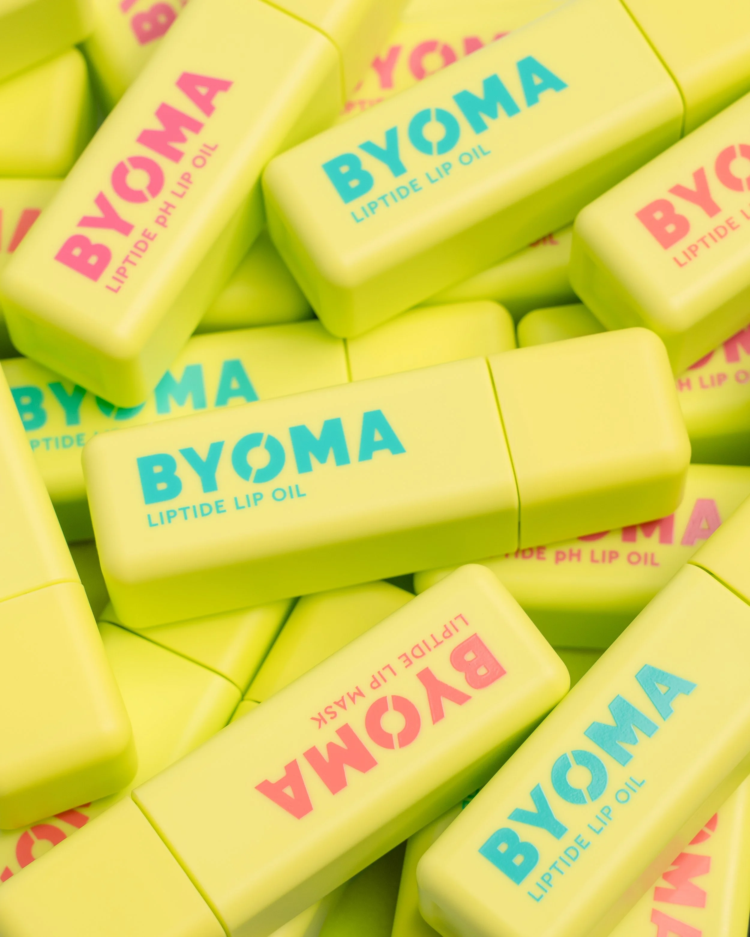 Several yellow lip balm containers labeled BYOMA LIPTIDE LIP OIL in pink and blue text, scattered and stacked together.