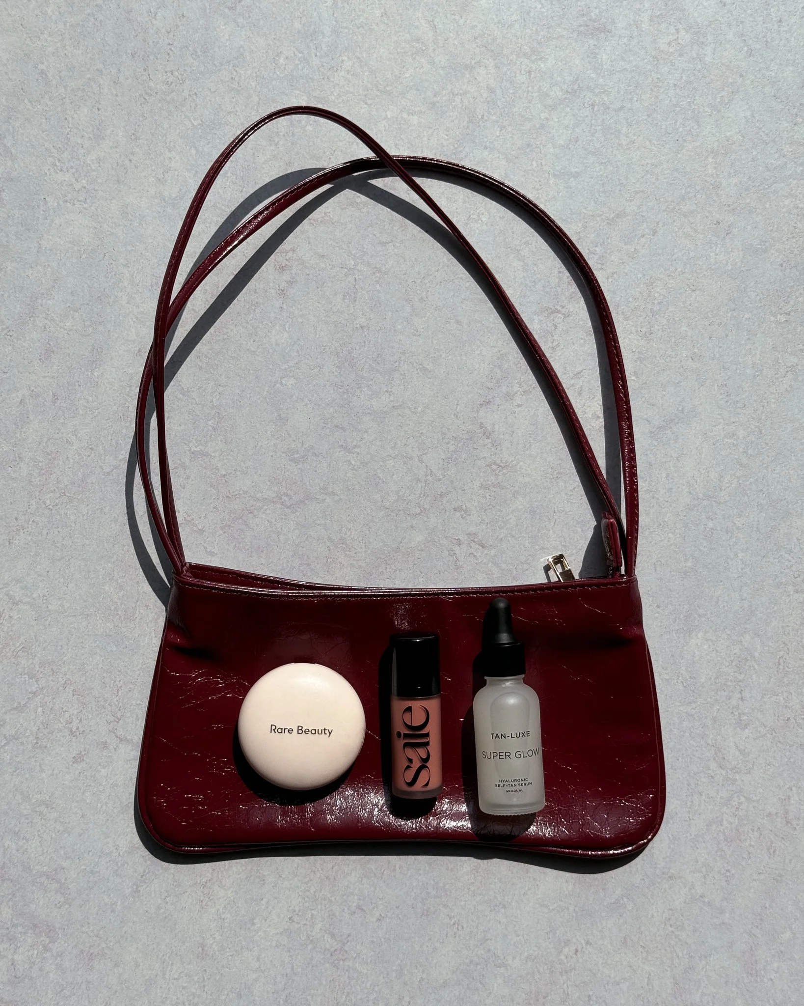 Red handbag with three beauty products inside: a white compact labeled 'Rare Beauty,' a small black and pink bottle labeled 'Sailie,' and a gray bottle with a dropper labeled 'Tan-Luxe Super Glow' on a light gray surface.