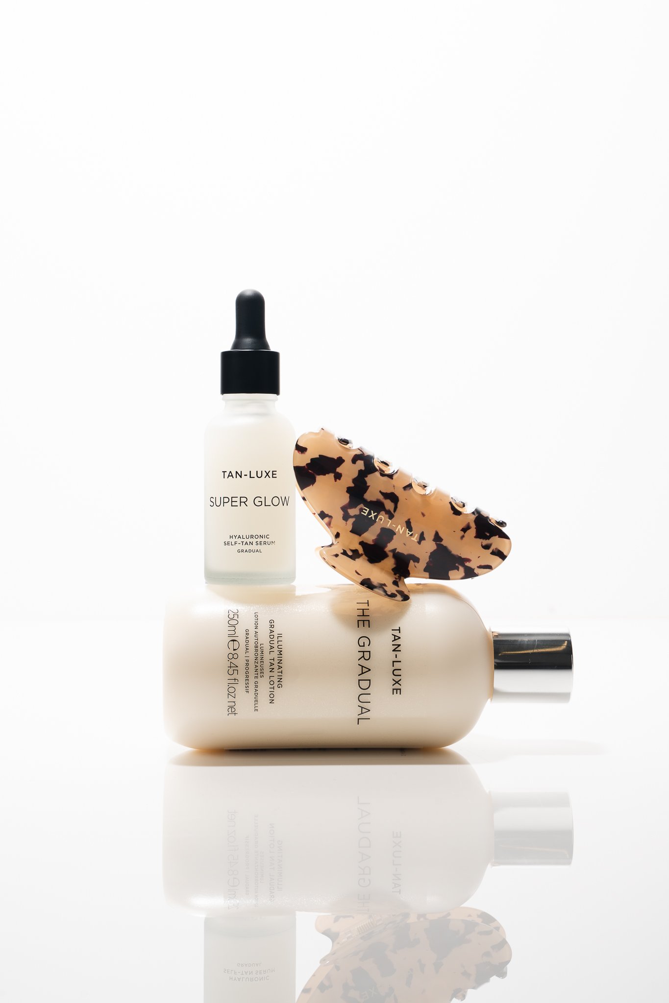 Skincare products including a bottle of Tan-Luxe Super Glow serum, The Ordinary illuminating serum, and a tortoise shell hair clip with a cream background.