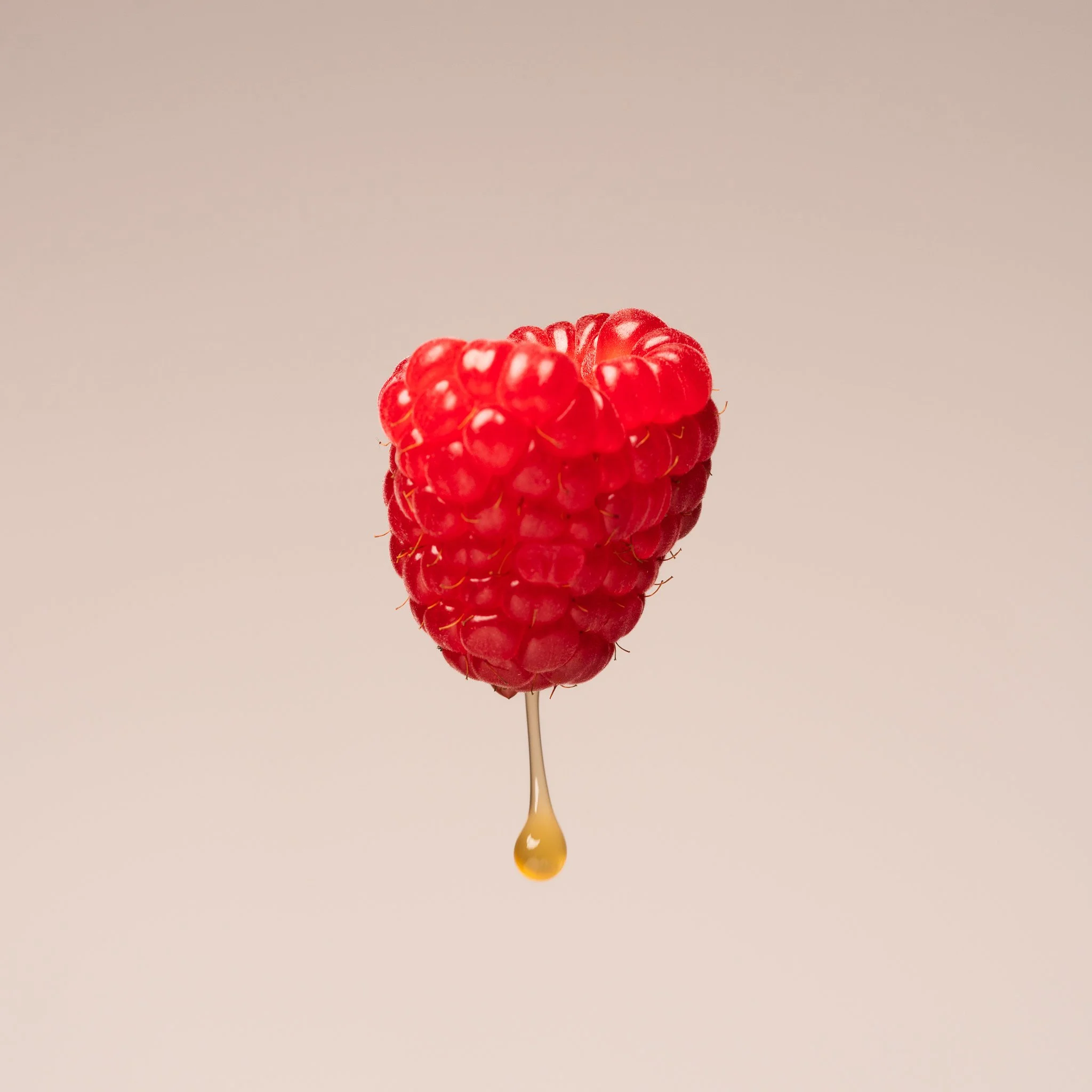 A close-up of a raspberry with a drop of honey hanging from its bottom.