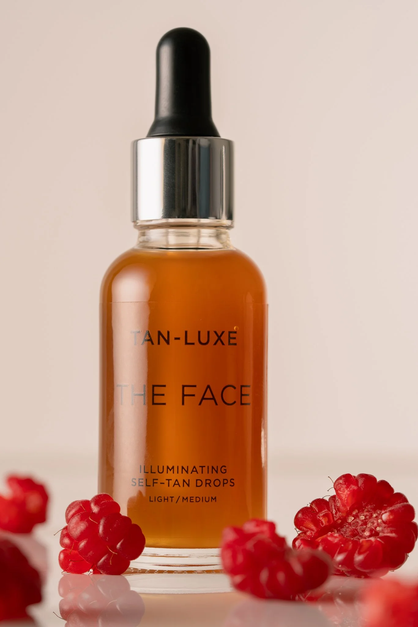 A bottle of Tan-Luxe The Face illuminating self-tan drops in front of raspberries on a neutral background.