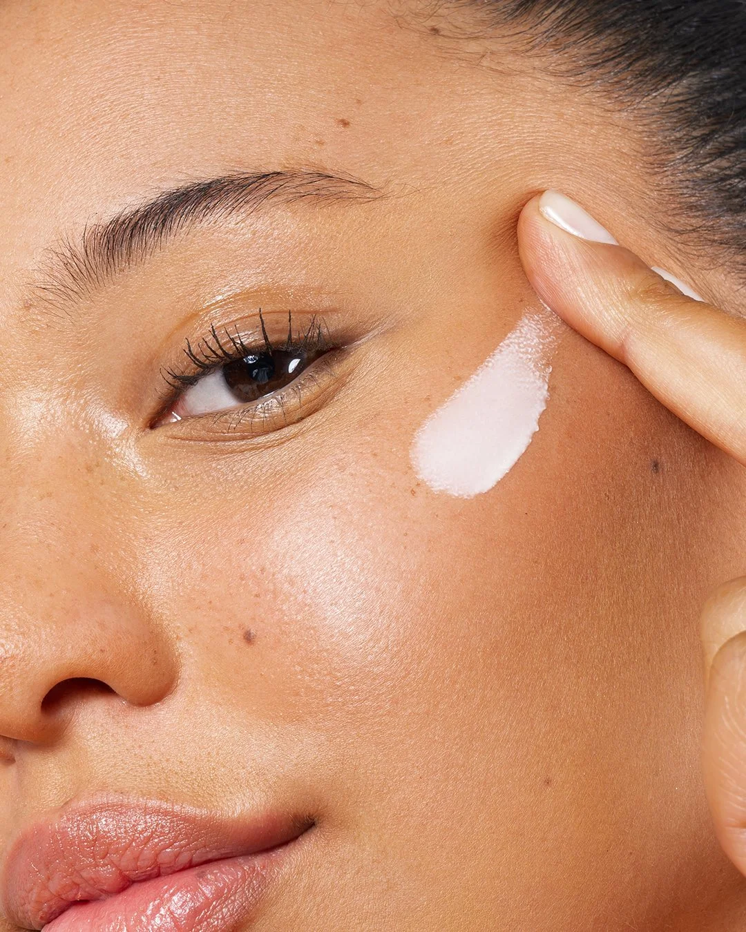 Close-up of a woman's face with a dab of white cream under her eye, as she points to it with her finger.