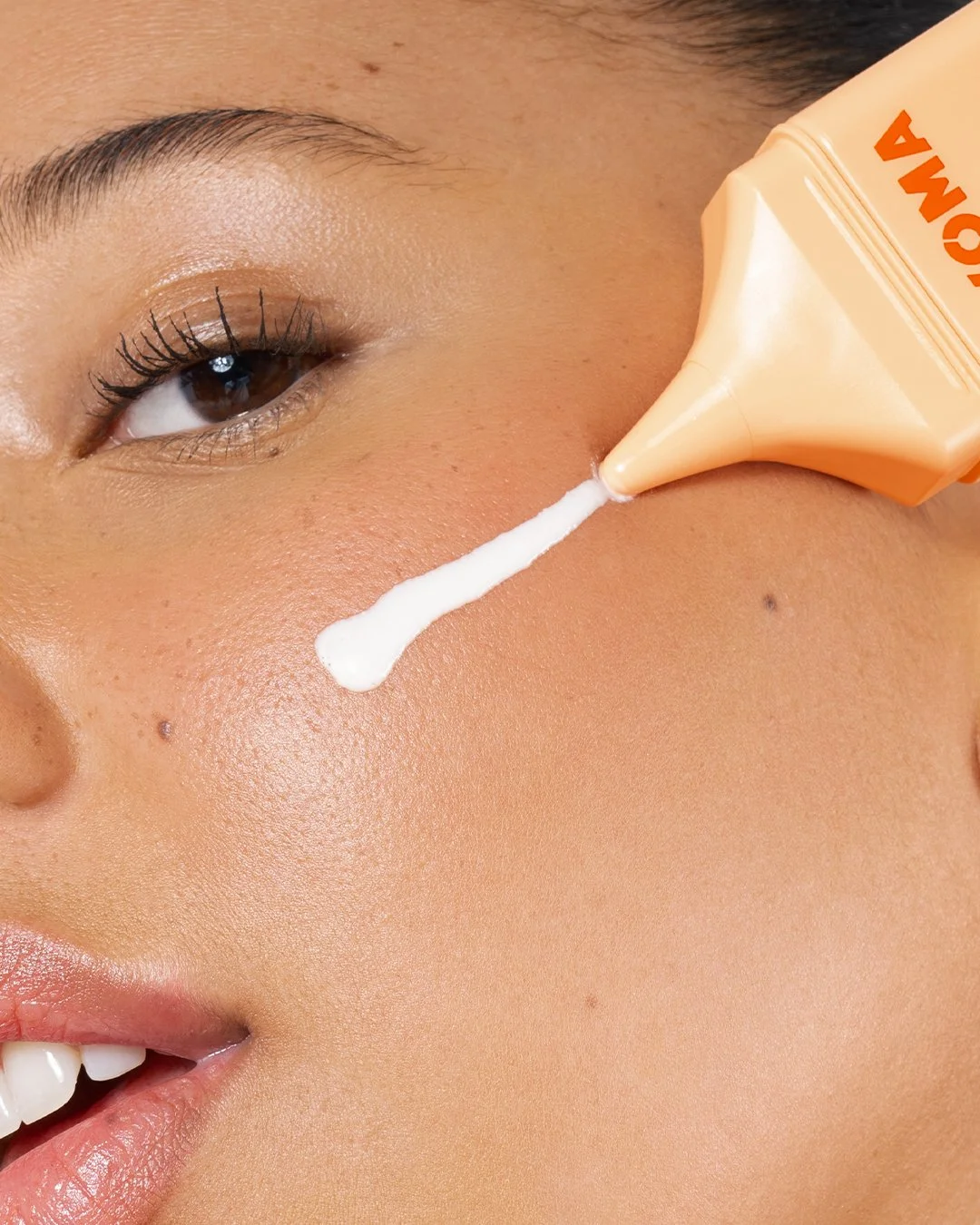 Close-up of a person applying white cream from an orange squeeze bottle onto their cheek. The person's eye is partially open, showing the brown iris and long eyelashes. The skin appears smooth and glowing.