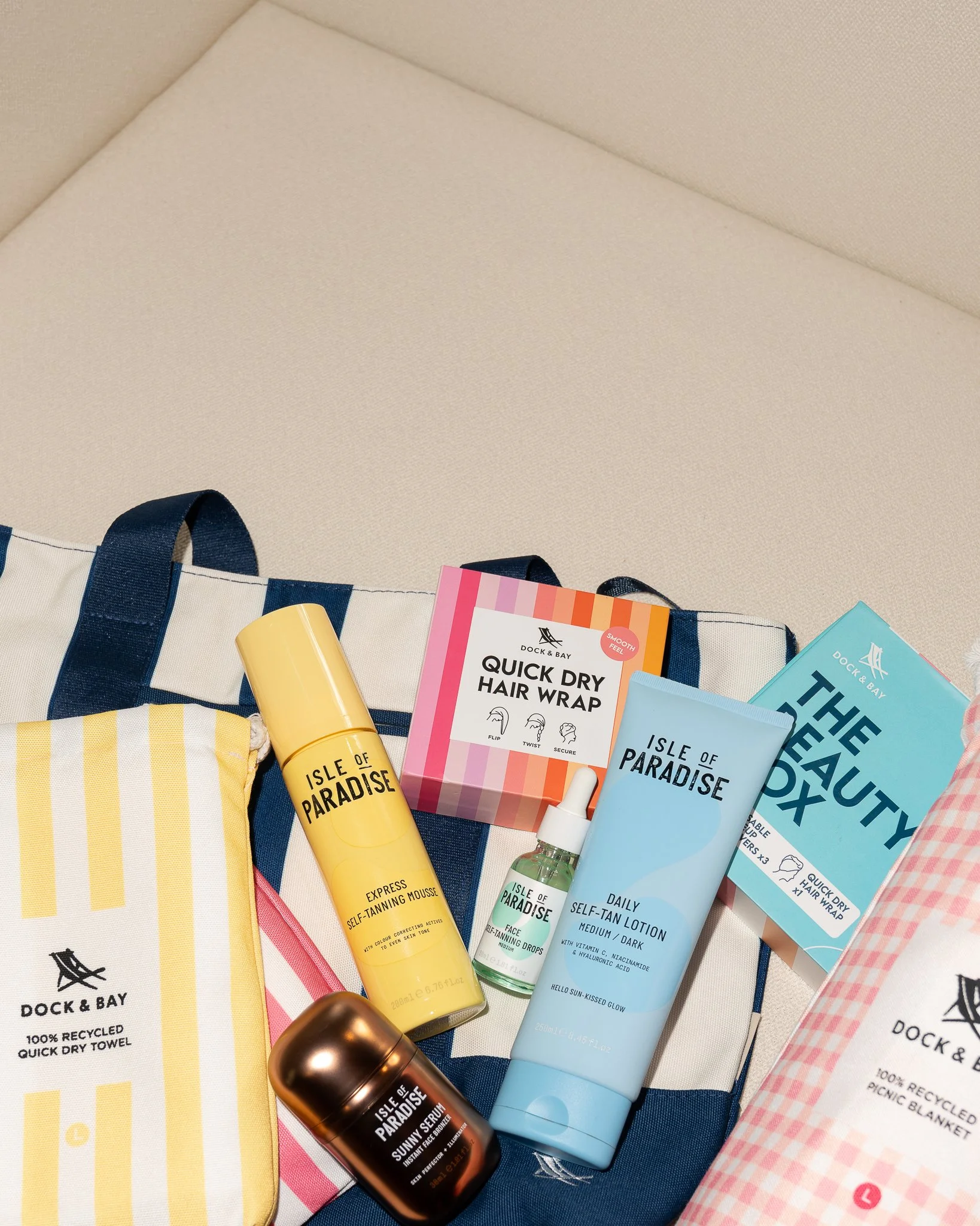Collection of skincare and beauty products, including self-tanning mousse, lotion, and face drops, arranged on a bag and towel with Beach theme.
