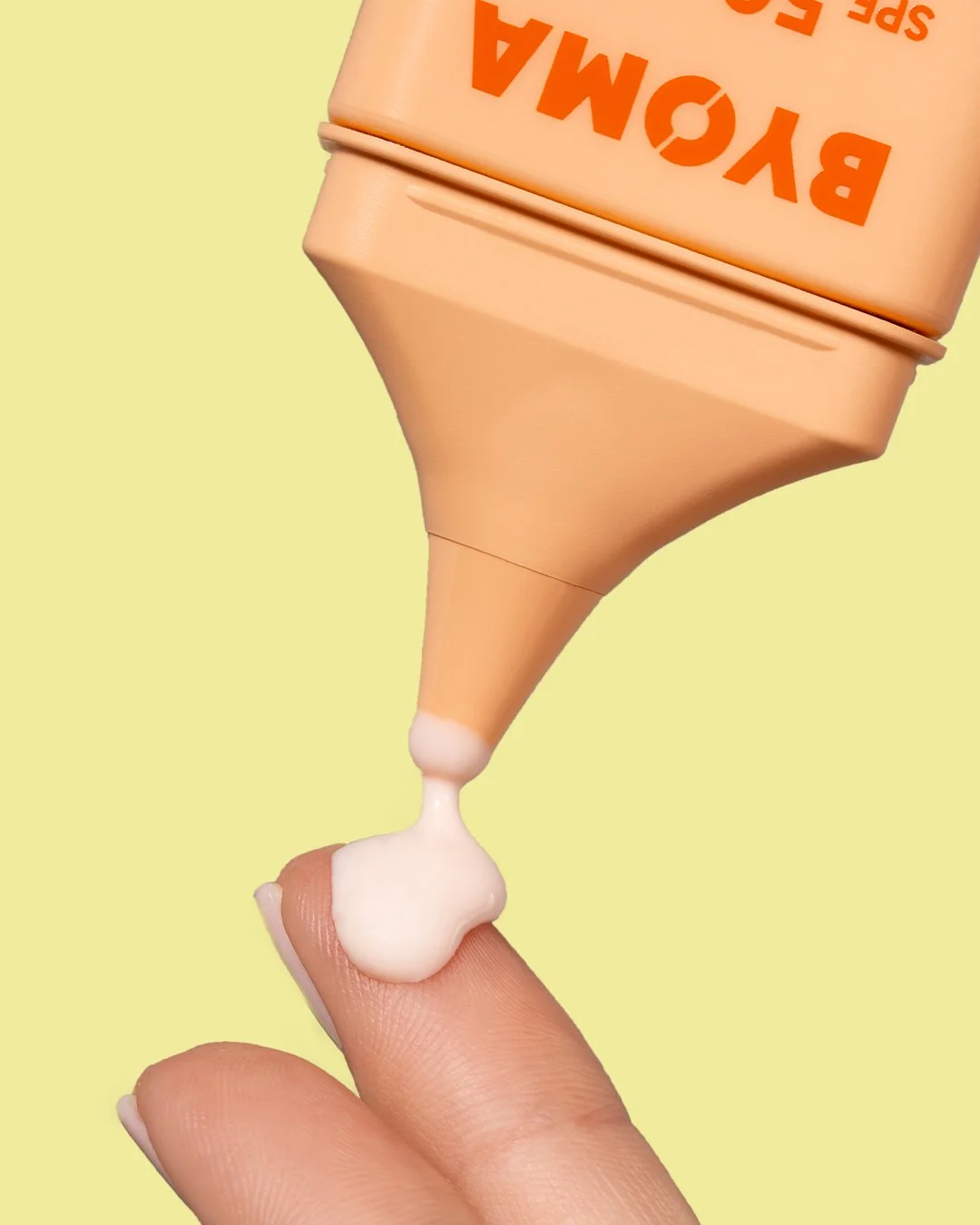A squeeze bottle of B YOMA SPF 50 sunscreen being used to apply lotion on a person's finger, with a light yellow background.