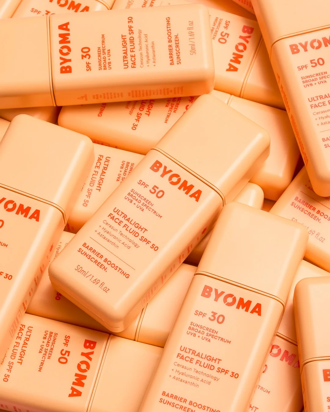 Multiple bottles of BYOMA Ultra Lightweight Face Fluid SPF 30 sunscreen with orange text on beige background, overlapping and stacked.
