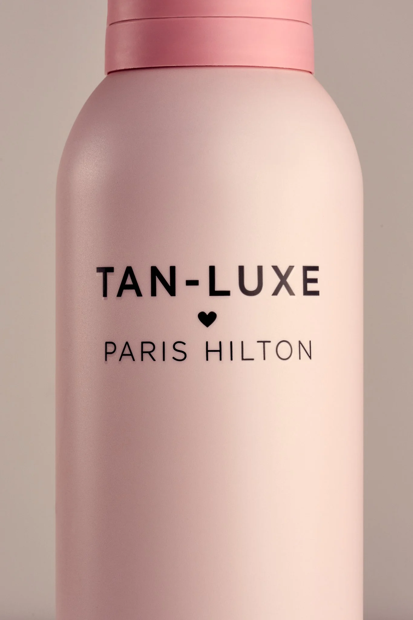 A pink bottle with black text reading "TAN-LUXE PARIS HILTON" and a small black heart symbol in the center.