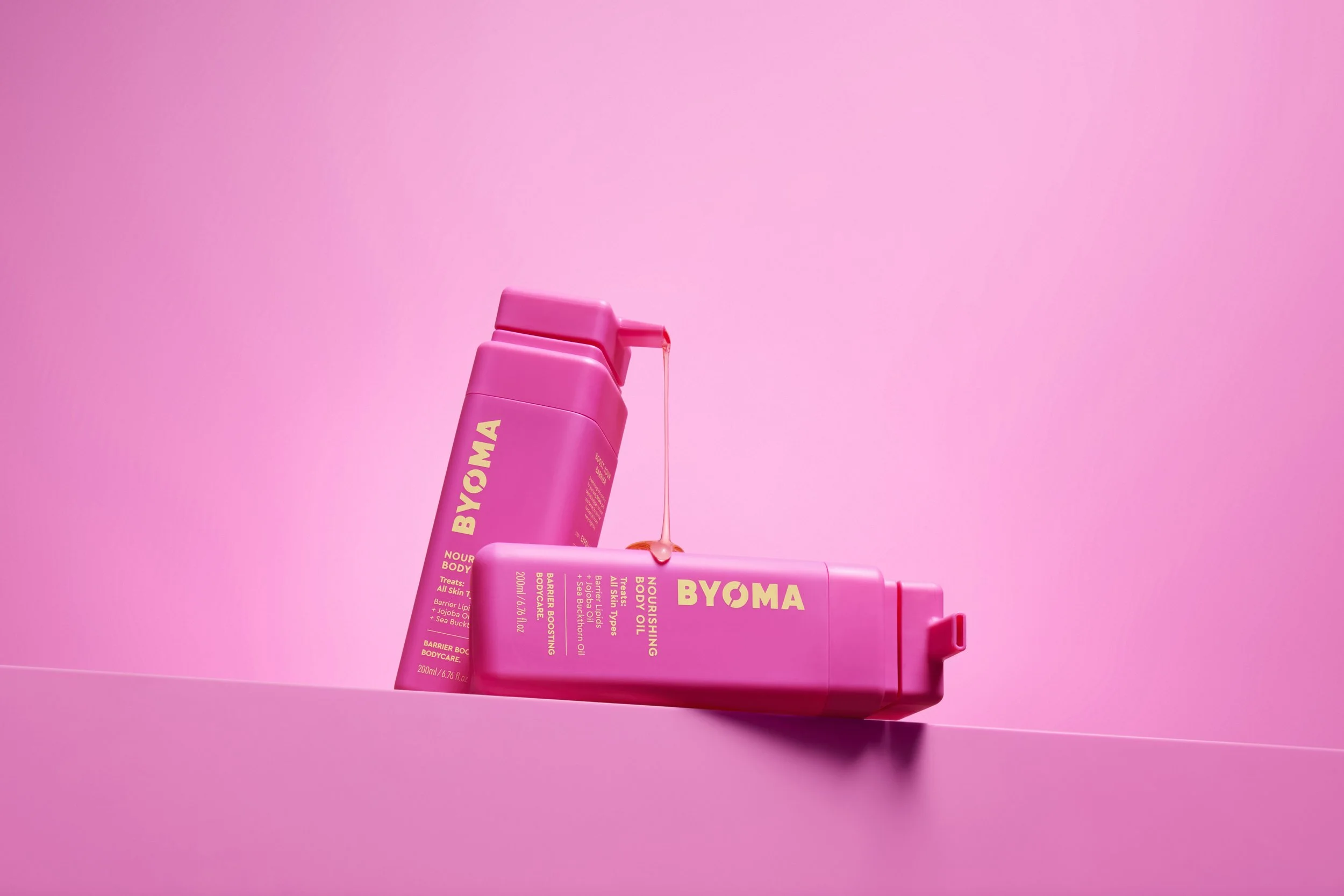 Two pink bottles of BYOMA skincare products, one with a cap open and pink liquid dripping from it, against a pink background.