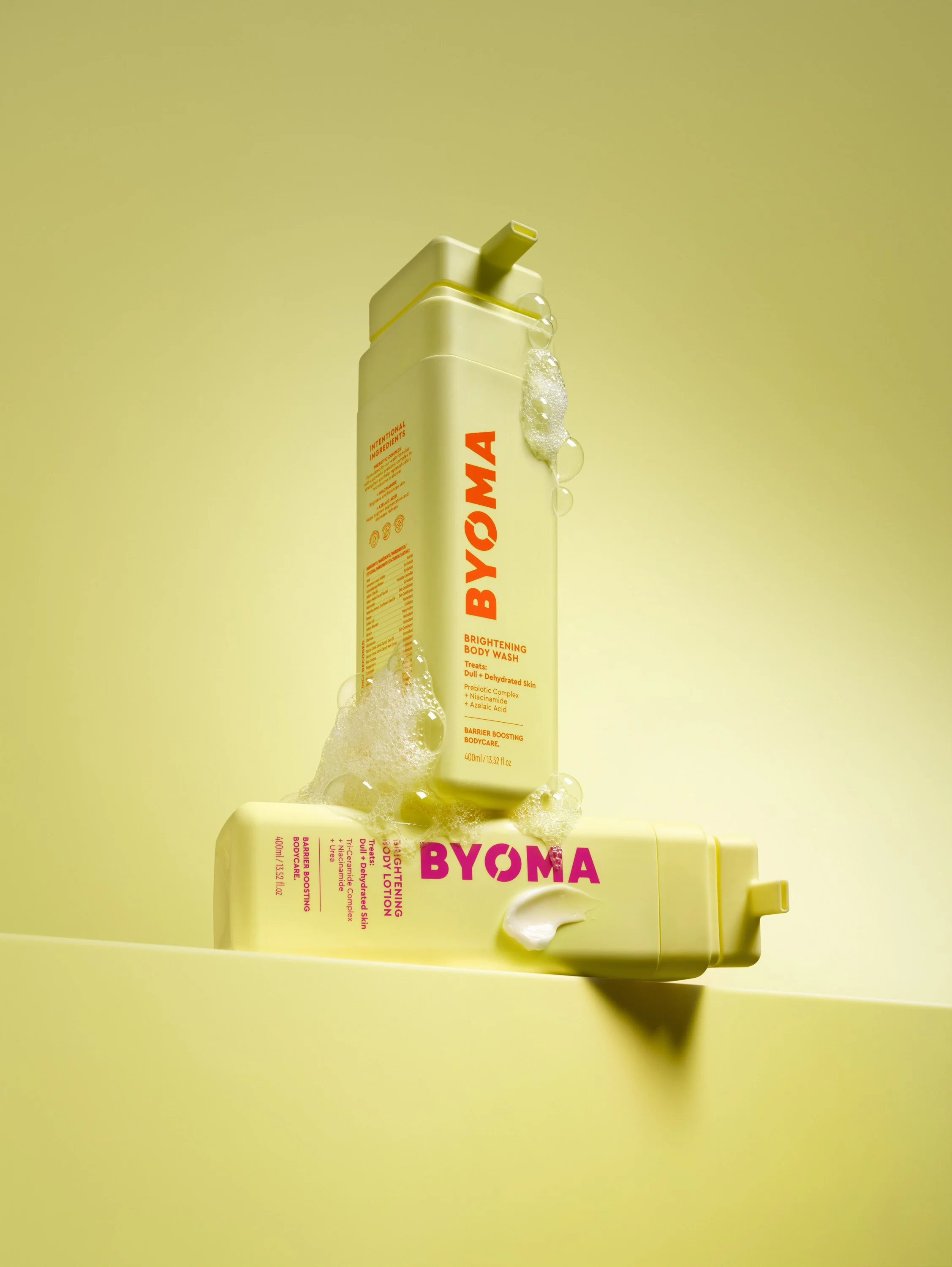 Two yellow bottles of BYOMA skincare products, one labeled brightening body wash and the other brightening body lotion, with foam and bubbles on the bottles against a yellow background.