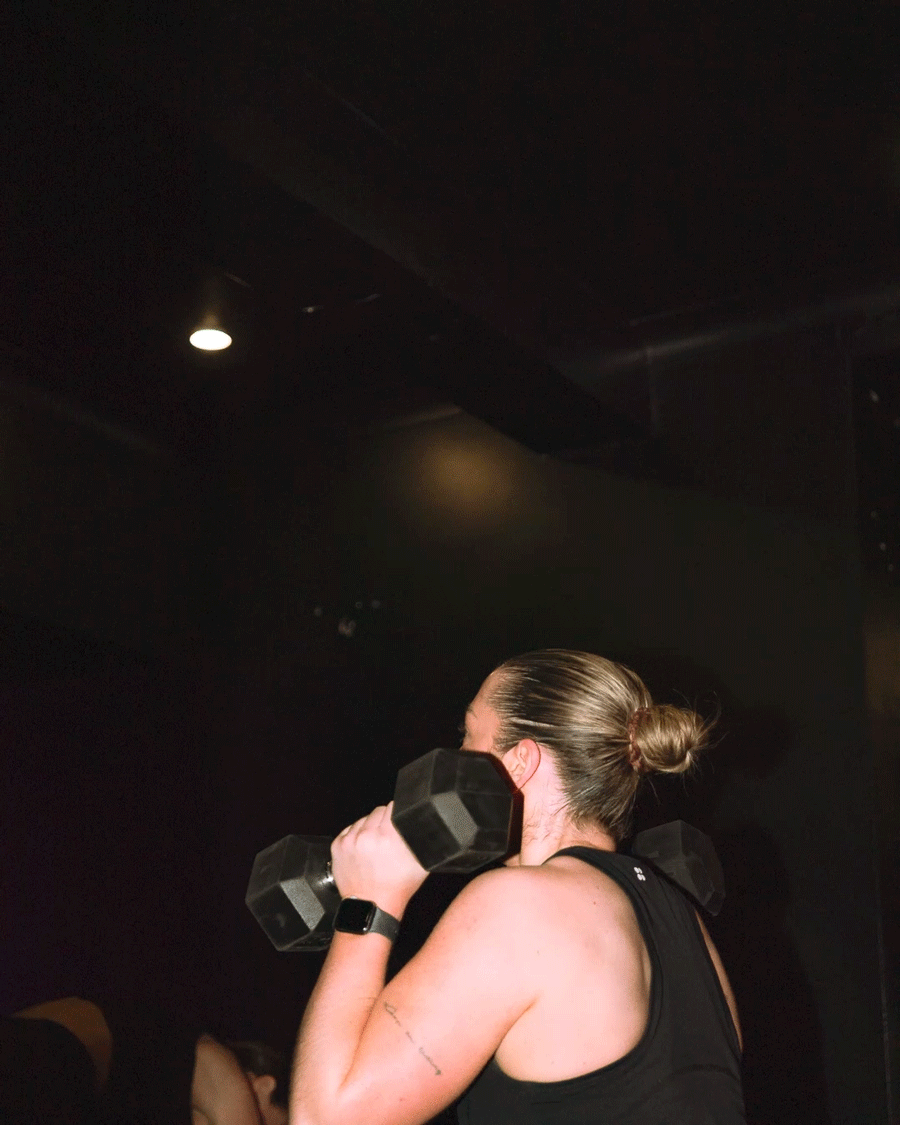 Woman with a bun hairstyle, wearing a black tank top, lifting black dumbbells in a dark gym.