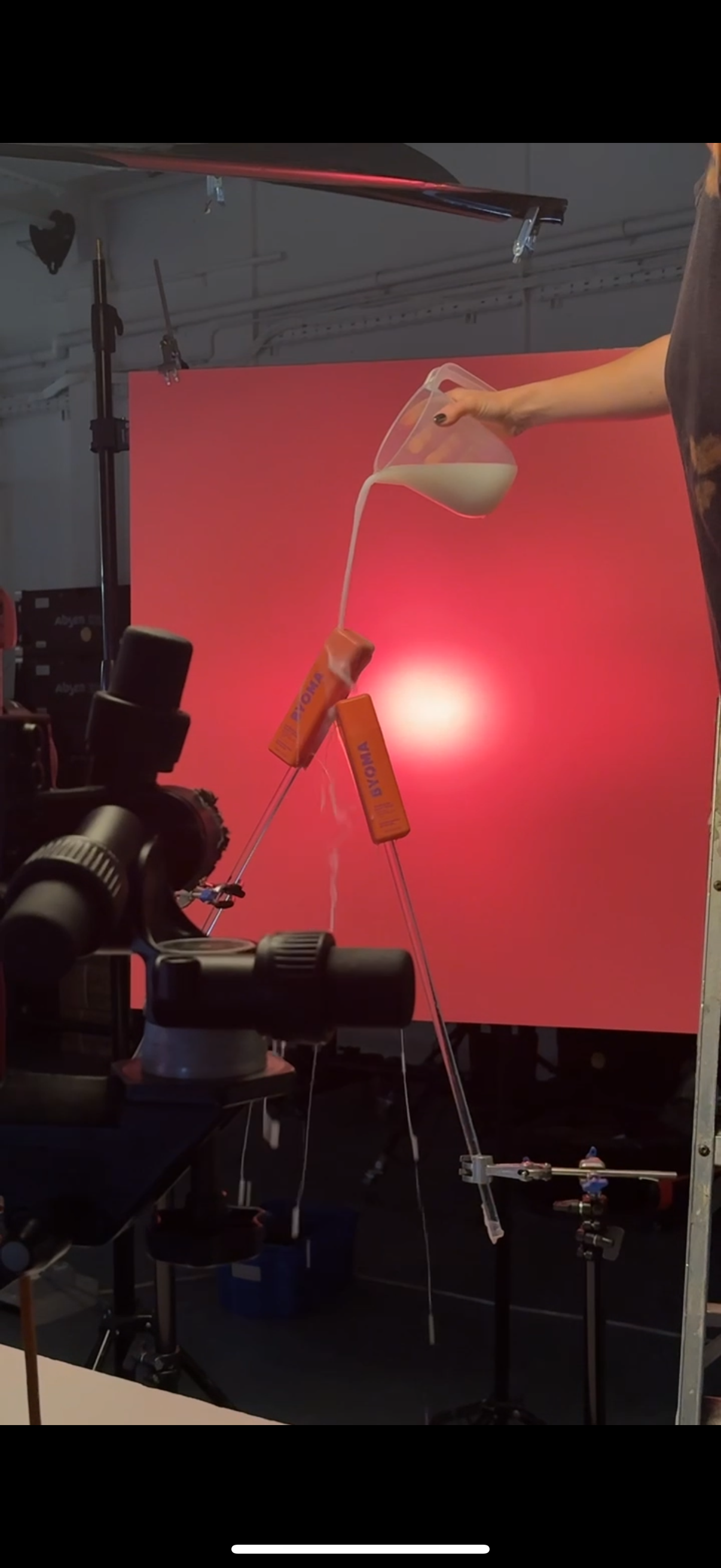 A person pours liquid from a clear measuring cup into an orange device mounted on a tripod, with a pink background and studio lighting setup.