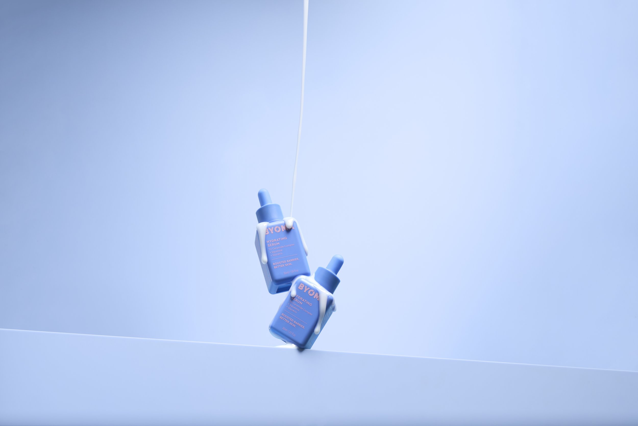 Two blue bottles of BYOMA hydrating serum with white caps and drips of serum on the bottles, hanging in mid-air against a light blue background.