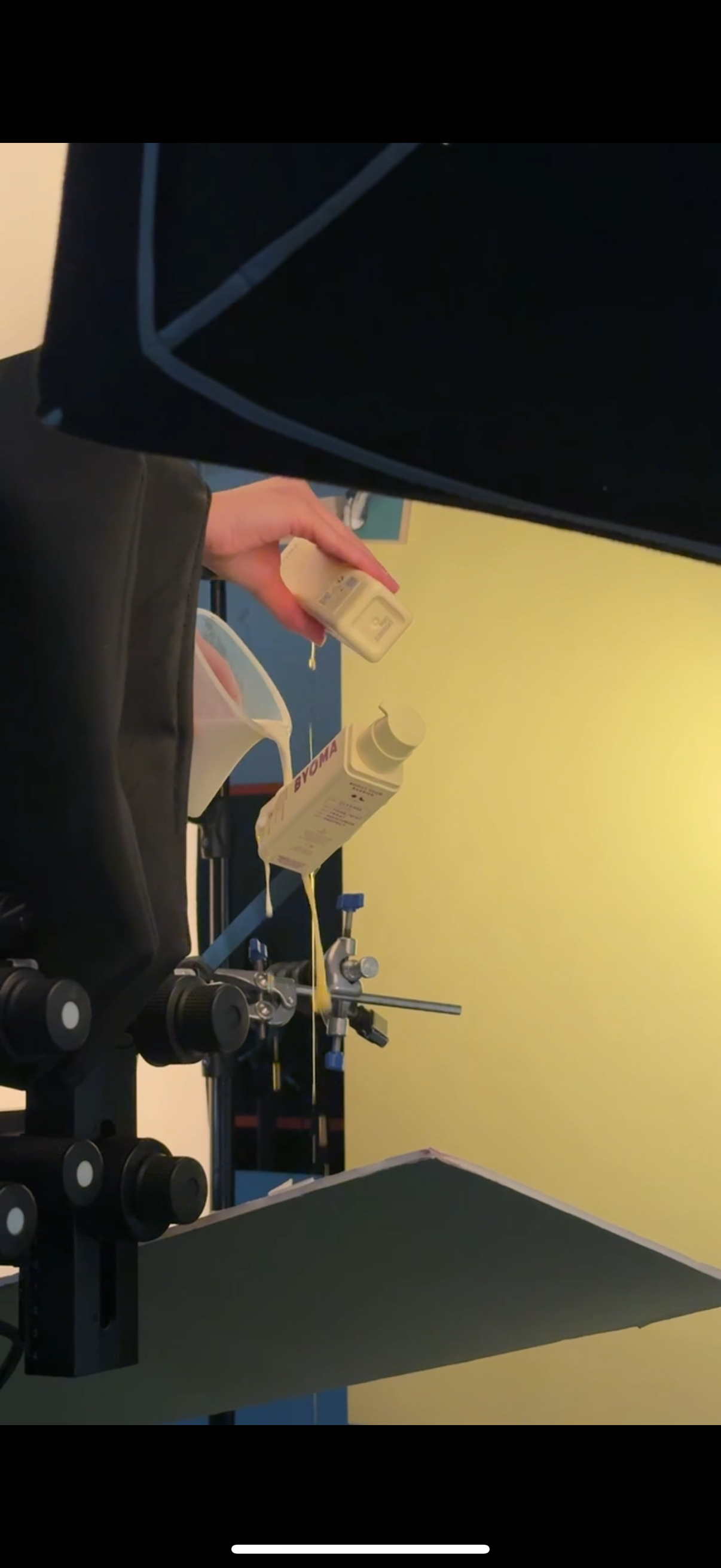 A person pouring yellow liquid medicine from a bottle into a medical device with tubes, on a black stand, with a yellow wall in the background.