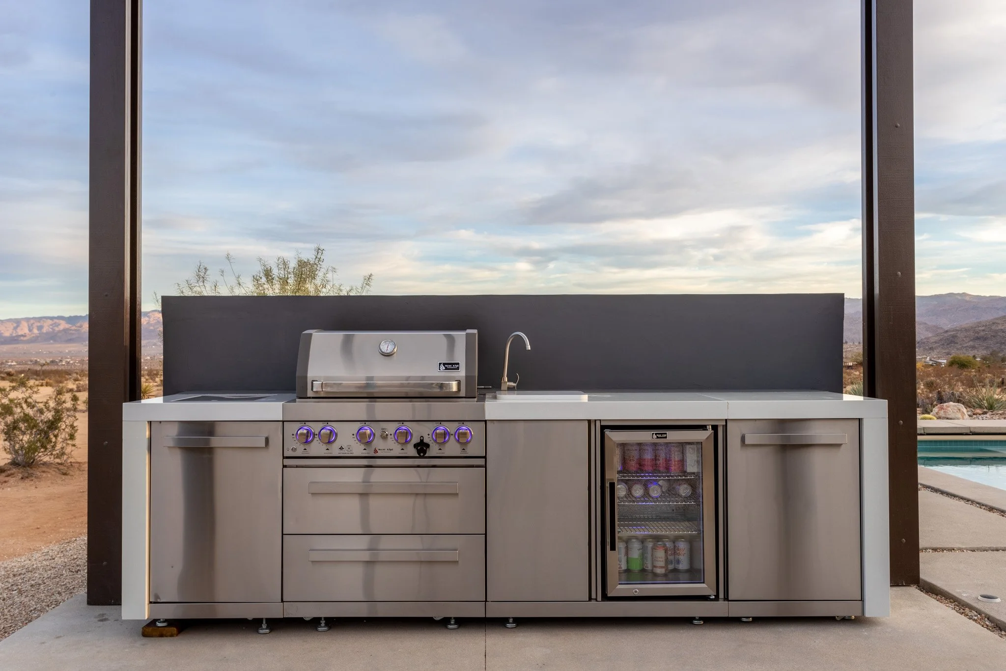 Outdoor kitchen with stainless steel grill, mini refrigerator, and sink, set against a desert landscape with mountains and a cloudy sky.