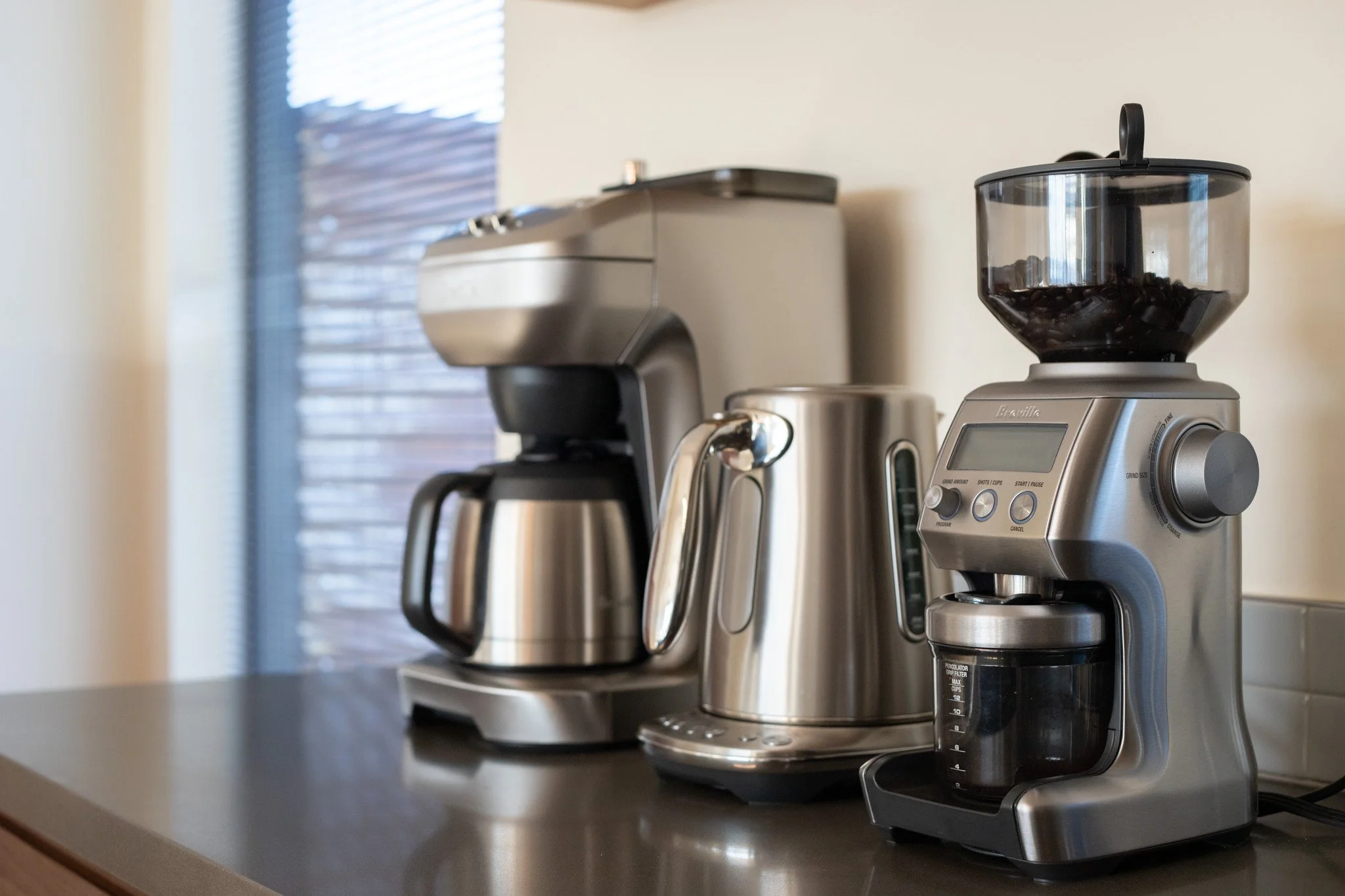 Coffee maker, electric kettle, and coffee grinder on kitchen counter with a window in the background.