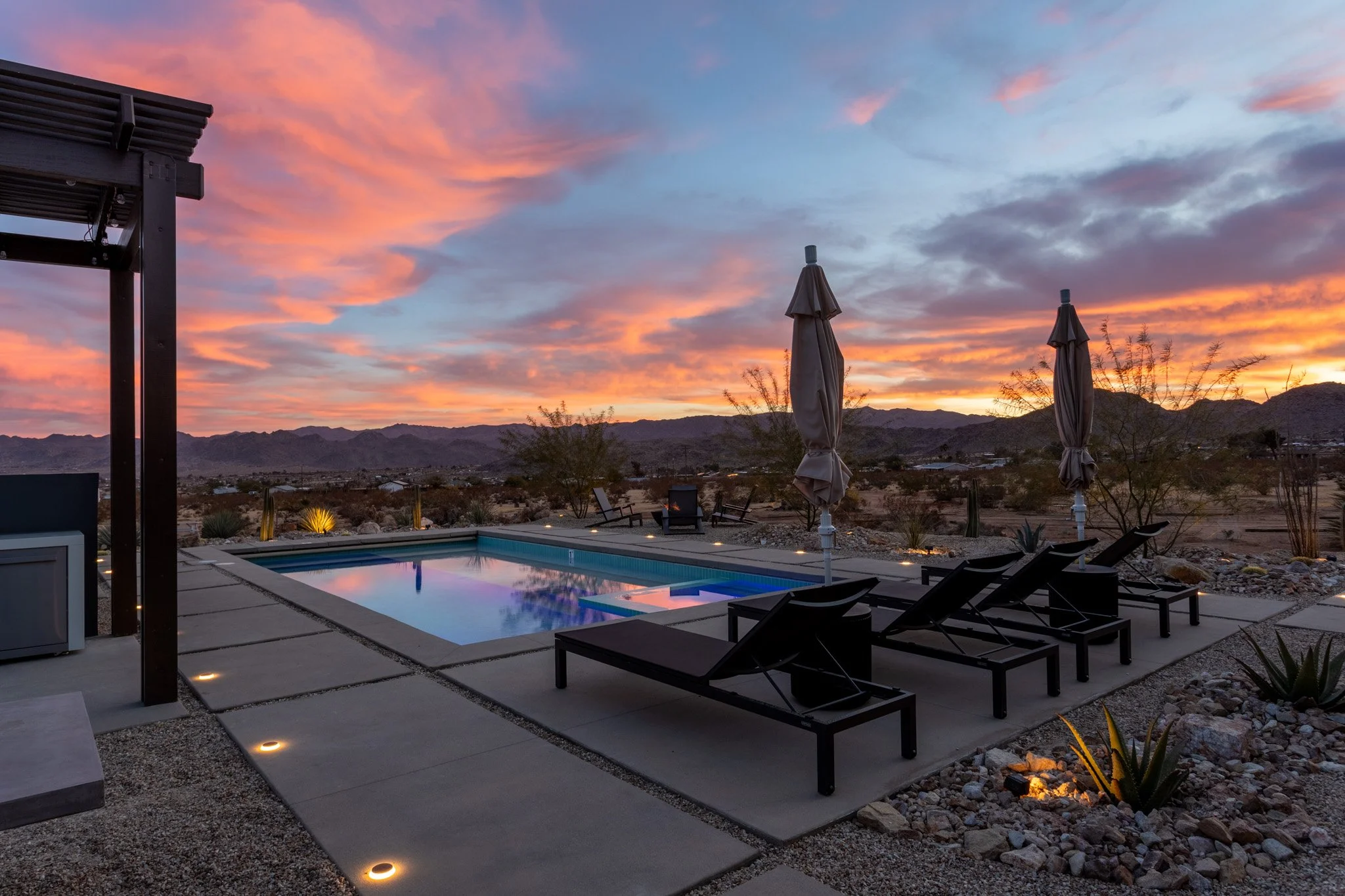 A desert backyard with a swimming pool at sunset, lounge chairs, closed umbrellas, and mountain silhouettes in the distance.