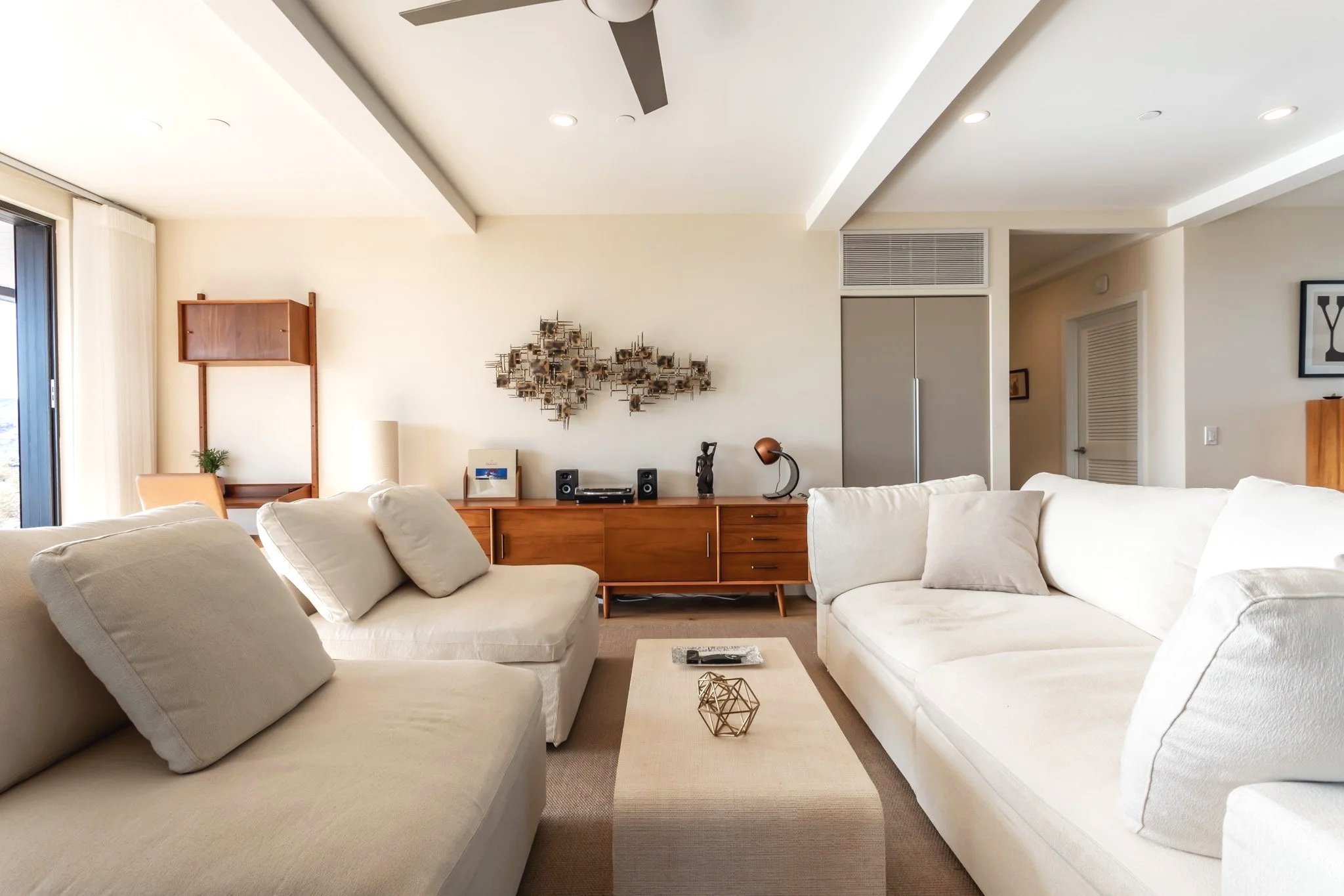 Bright living room with white sofas, a wooden sideboard, and contemporary decor, including wall art and sculptures.