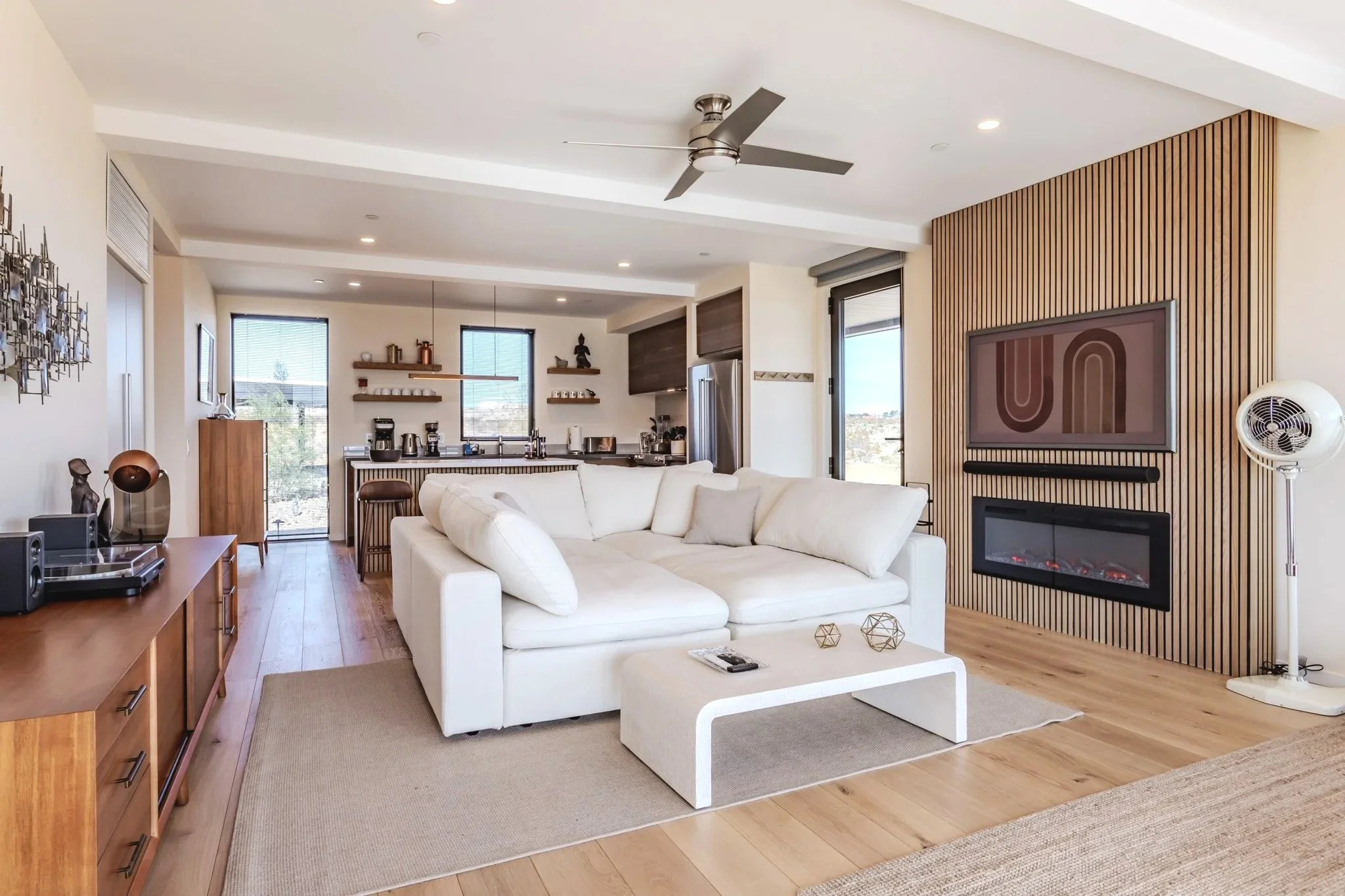 Modern living room with white sofa, wooden flooring, wall-mounted TV, and a fireplace, open to a kitchen area with windows and barstools.