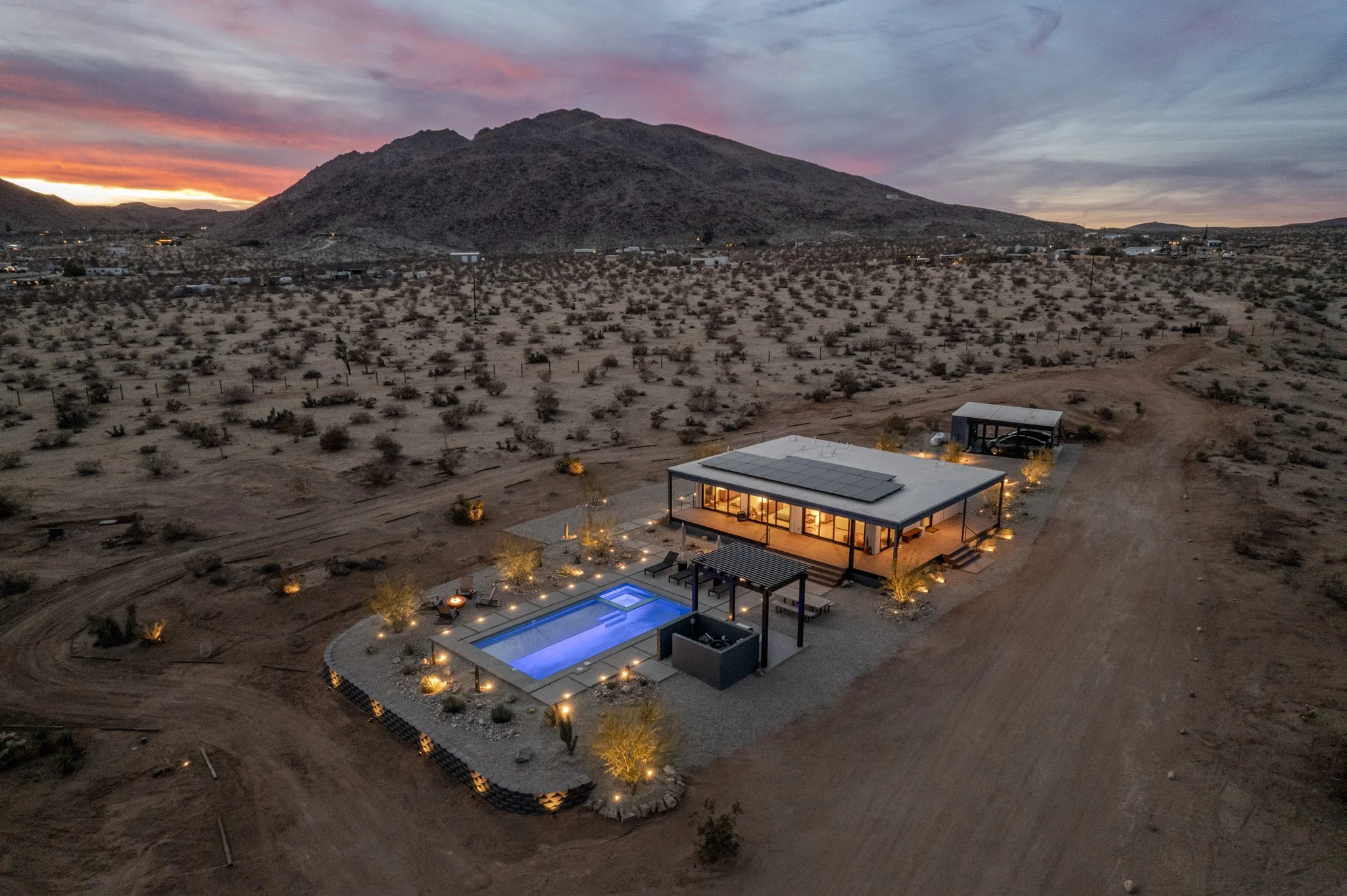 A modern house with solar panels on the roof, surrounded by desert terrain, a swimming pool with blue lighting, outdoor seating, and decorative lighting at sunset with a mountain in the background.